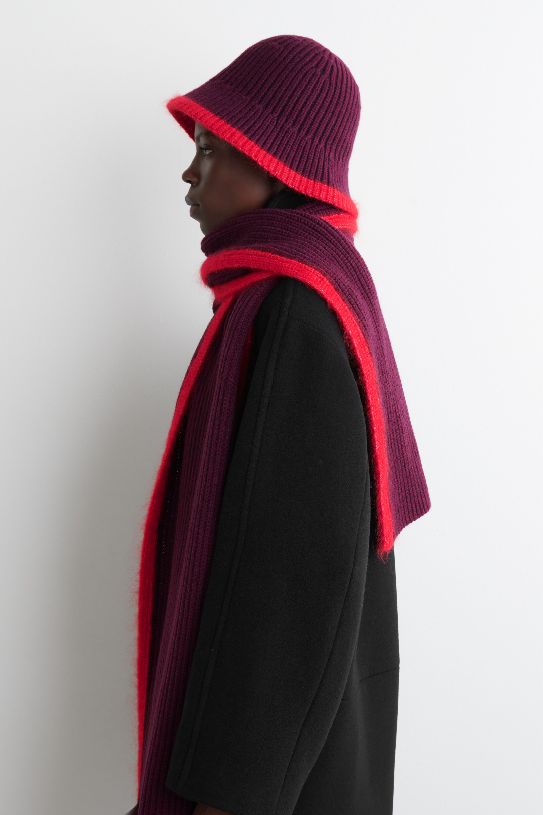 Agrandir l'image: Un ensemble de bonnet et écharpe tricotés est présenté de profil gauche. Le chapeau violet foncé côtelé de style seau présente un bord rouge vif, et l'écharpe assortie violette foncée côtelée, avec des rayures rouges vives et une texture duveteuse, est enroulée autour du cou par-dessus un manteau noir.