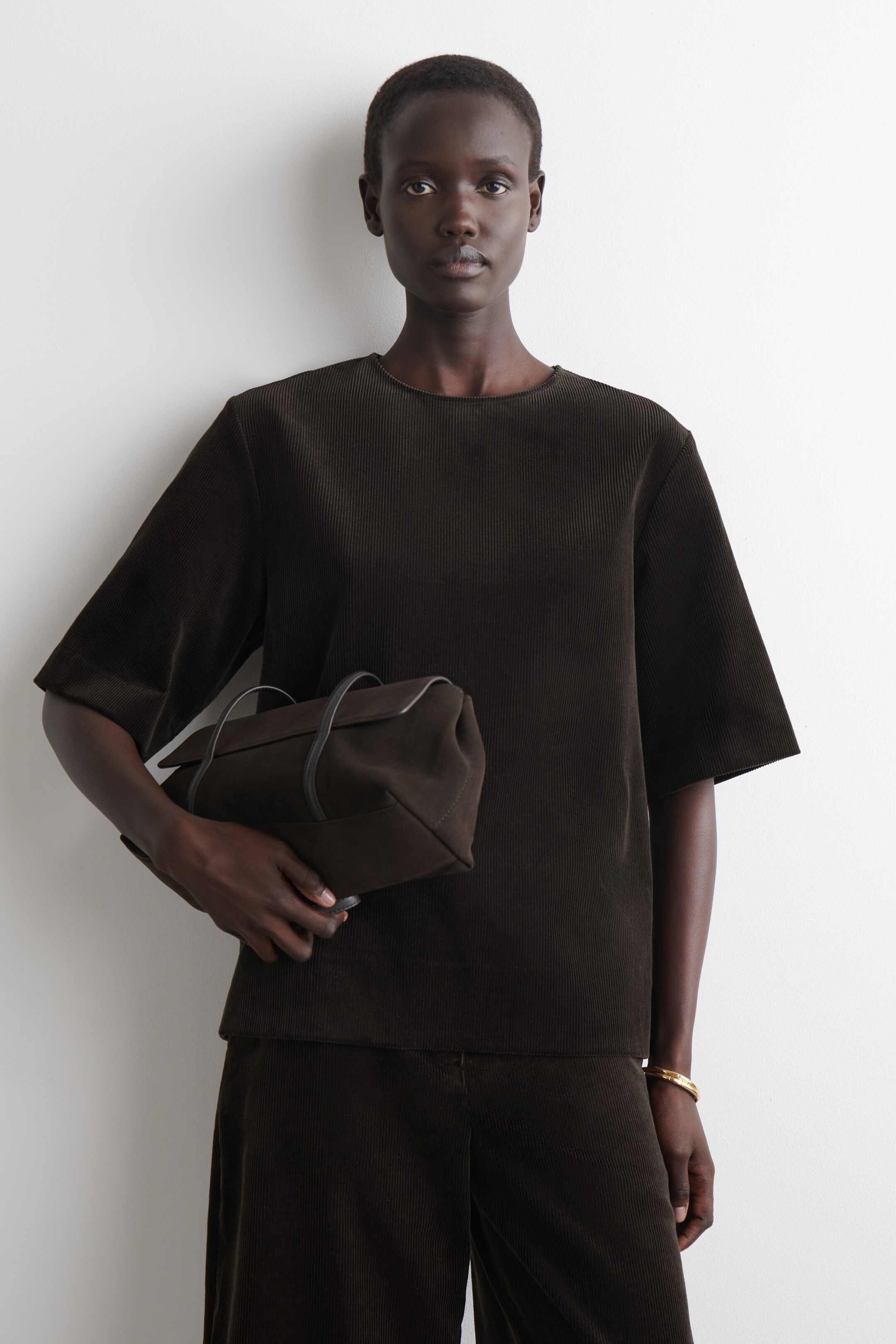 Visa större bild: En mörkbrun, finribbad topp med rund hals och breda korta ärmar bärs tillsammans med matchande mörkbruna ribbade byxor med vida ben. En mjuk, rektangulär mörkbrun handväska med två korta svarta handtag hålls i ena handen.