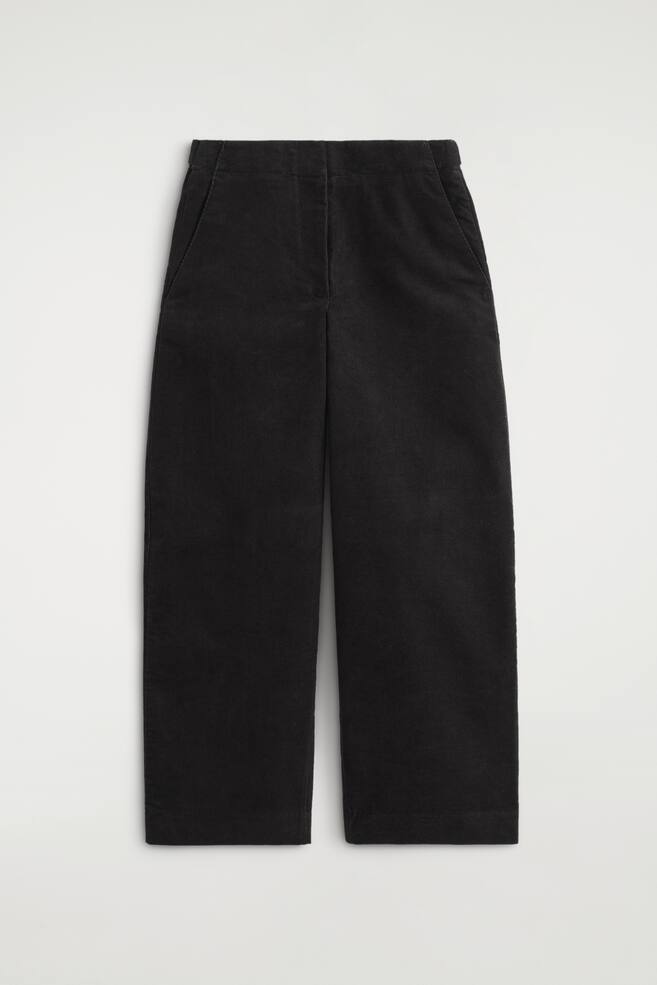 These black corduroy barrel-leg trousers are shown from the front, featuring a high-rise waistband, side pockets, and a clean front fly. The trousers display a structured, wide-leg silhouette that curves slightly outward and tapers towards the hem. - 1