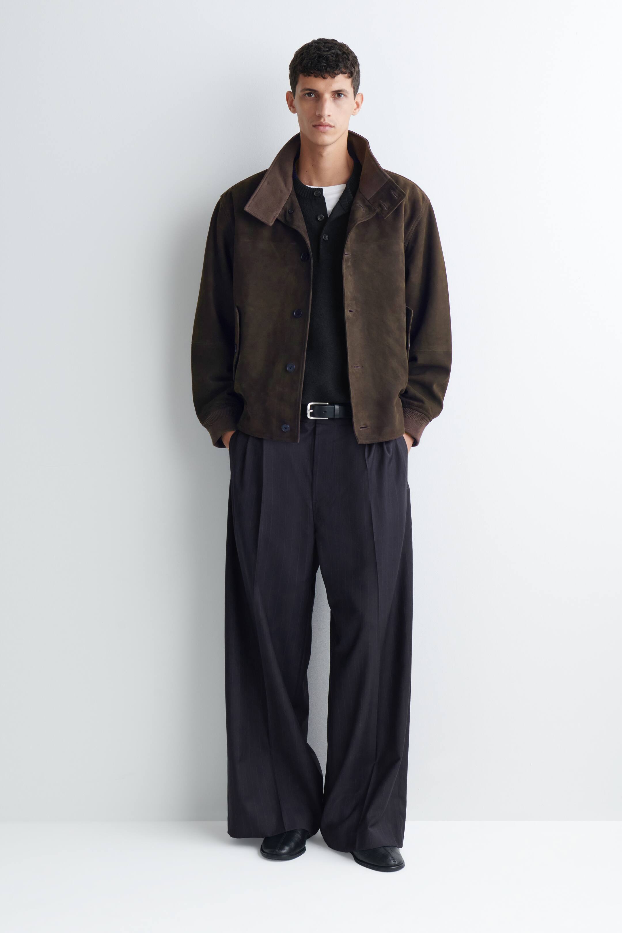 Agrandir l'image: Un pantalon plissé à coupe décontractée et jambe large, en gris foncé, est porté par le mannequin, et est associé à une ceinture en cuir noire à la taille. Le pantalon présente de profondes pinces sur le devant et une silhouette ample et fluide, qui retombe légèrement sur des chaussures en cuir noir.