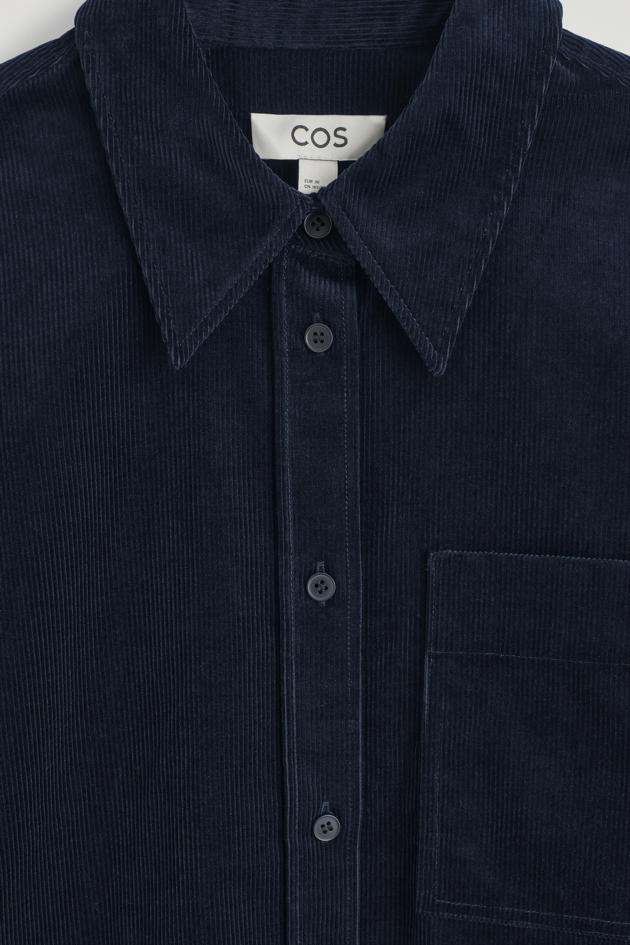 A close-up front view showcases a navy blue cotton corduroy shirt with a fine ribbed texture. The shirt features a pointed collar, a button-down placket with dark buttons, and a single chest patch pocket. A close-up front view showcases a navy blue cotton corduroy shirt with a fine ribbed texture. The shirt features a pointed collar, a button-down placket with dark buttons, and a single chest patch pocket.