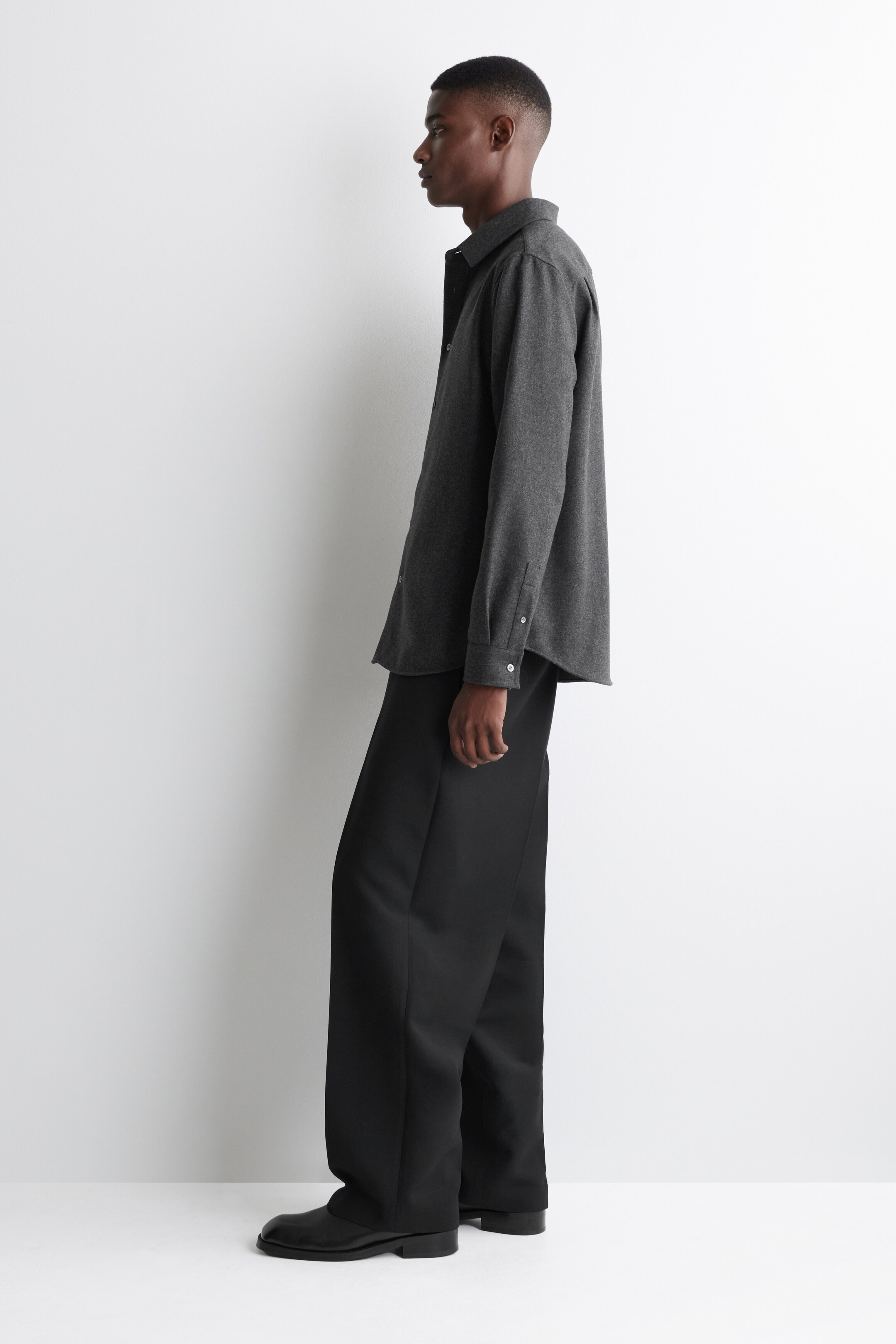 Visualizza immagine più grande: Una camicia a maniche lunghe grigio scuro e pantaloni a gamba larga neri sono presentati di profilo. La camicia a vestibilità rilassata è rifinita con un colletto a punta e polsini abbottonati, mentre i pantaloni a gamba larga cadono morbidi su scarpe nere.
