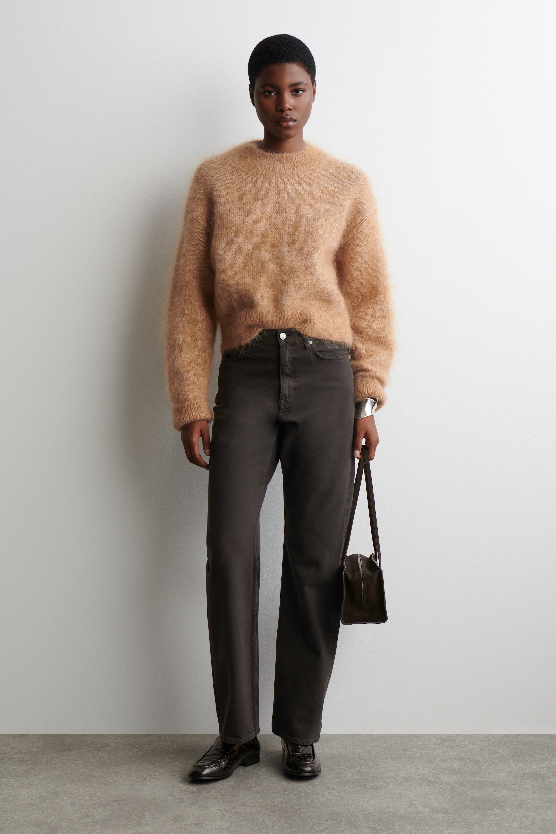 Ver imagen más grande: Un jersey de cuello redondo marrón claro y peludo se lleva en un ajuste relajado, combinado con pantalones rectos de pierna recta de color gris oscuro que descansan en la cintura natural. Dark brown textured loafers are being worn on the feet, and a dark brown small bag is being held at the side, all presented from a front view.