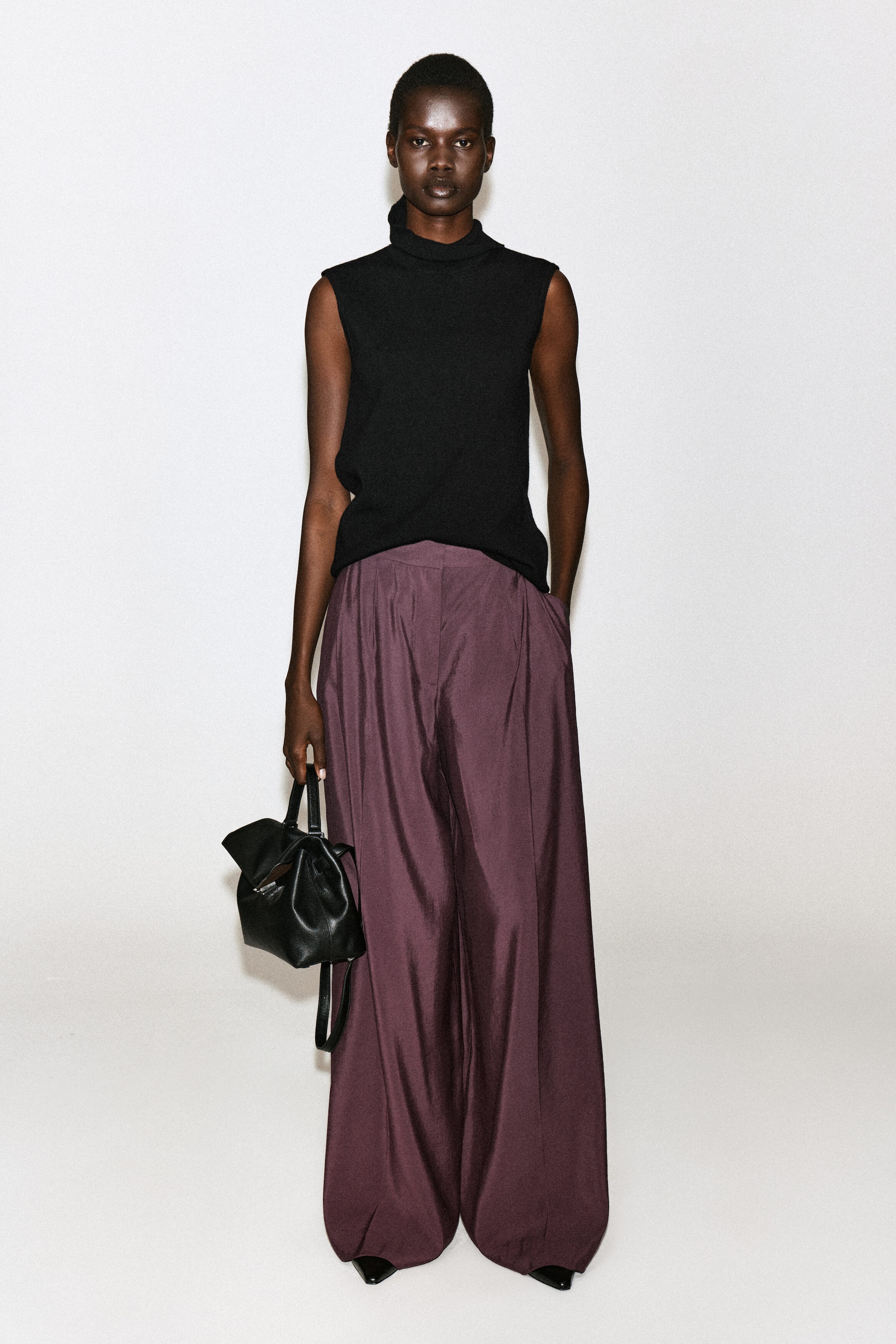 Gefaltete Wide-Leg-Hose aus Modal in tiefem Pflaumenton, vom Model getragen und mit einem schwarzen ärmellosen Rollkragen-Top kombiniert. Die Hose verfügt über eine lässige, fließende Silhouette mit Vorderfalten, die das Model aufgreift, indem es eine Hand in die Seitentasche steckt und dabei eine schwarze Ledertasche trägt. Gefaltete Wide-Leg-Hose aus Modal in tiefem Pflaumenton, vom Model getragen und mit einem schwarzen ärmellosen Rollkragen-Top kombiniert. Die Hose verfügt über eine lässige, fließende Silhouette mit Vorderfalten, die das Model aufgreift, indem es eine Hand in die Seitentasche steckt und dabei eine schwarze Ledertasche trägt.