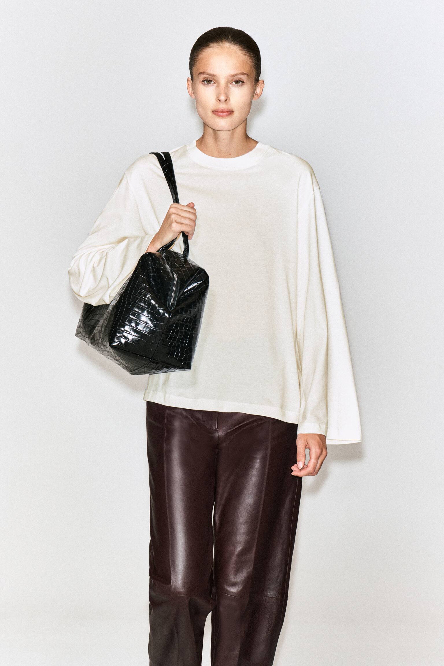 Una T-shirt a maniche lunghe in cotone spazzolato color bianco sporco Ã¨ indossata dalla modella, con vestibilitÃ  rilassata, girocollo a coste e spalle scese. La T-shirt Ã¨ abbinata a pantaloni in pelle marrone scuro, mentre la modella porta una borsa a mano nera effetto coccodrillo a tracolla su una spalla. - 1