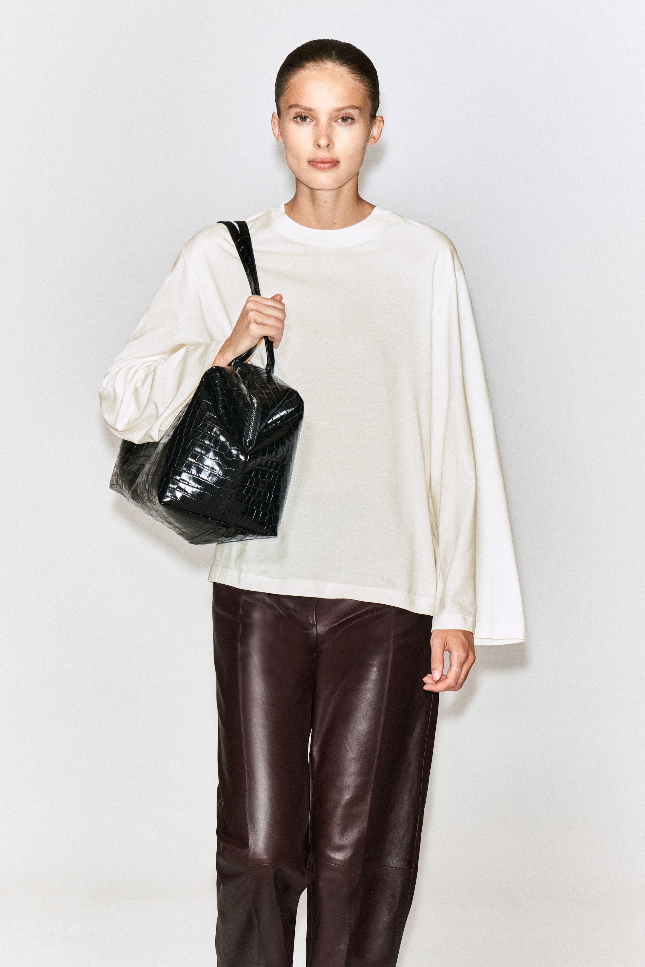 Visualizza immagine più grande: Una T-shirt a maniche lunghe in cotone spazzolato color bianco sporco è indossata dalla modella, con vestibilità rilassata, girocollo a coste e spalle scese. La T-shirt è abbinata a pantaloni in pelle marrone scuro, mentre la modella porta una borsa a mano nera effetto coccodrillo a tracolla su una spalla.