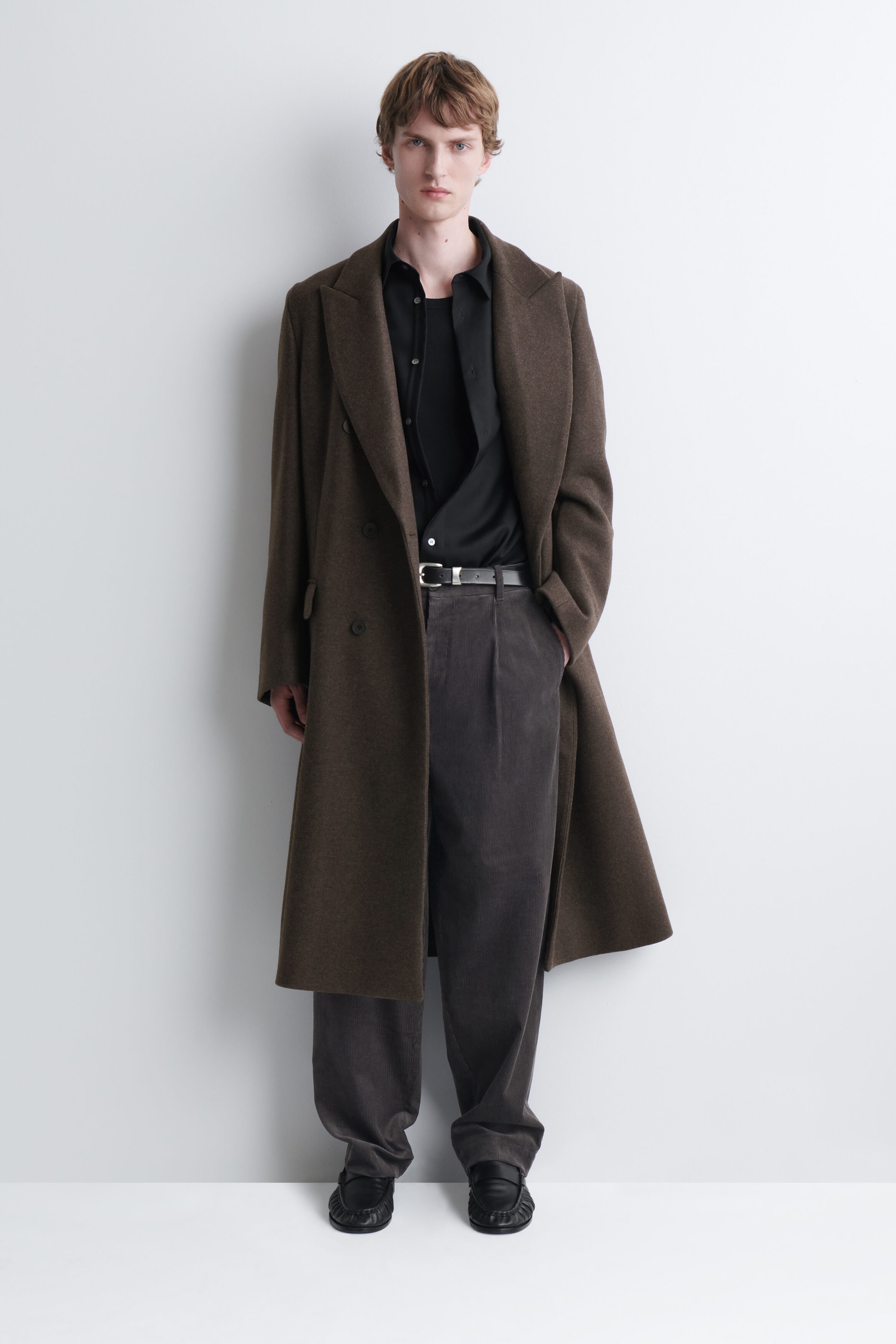 The model is wearing dark gray corduroy relaxed-fit tapered pants, styled with a black leather belt and a tucked-in black shirt. The pants feature subtle front pleats and a loose silhouette that tapers at the hem, layered under a long brown overcoat. The model is wearing dark gray corduroy relaxed-fit tapered pants, styled with a black leather belt and a tucked-in black shirt. The pants feature subtle front pleats and a loose silhouette that tapers at the hem, layered under a long brown overcoat.