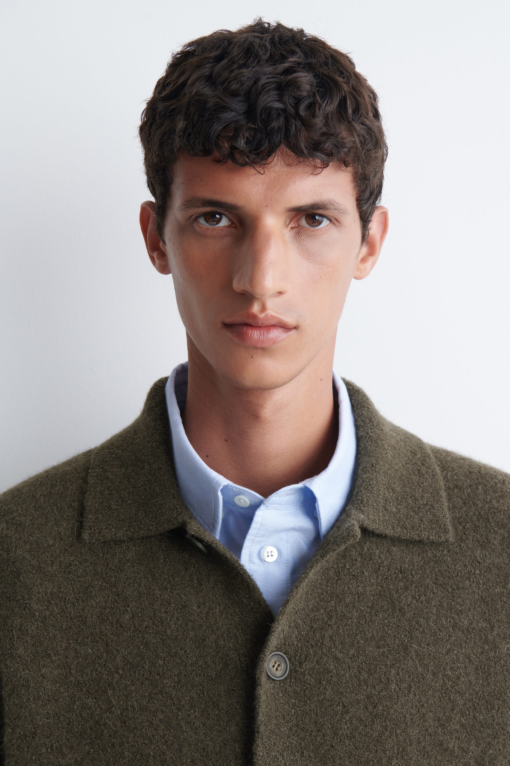 A textured collared cardigan in olive green is being worn by the model, layered over a light blue button-down shirt. The cardigan features a fuzzy, wool-like fabric, a flat collar, and front button closures. A textured collared cardigan in olive green is being worn by the model, layered over a light blue button-down shirt. The cardigan features a fuzzy, wool-like fabric, a flat collar, and front button closures.