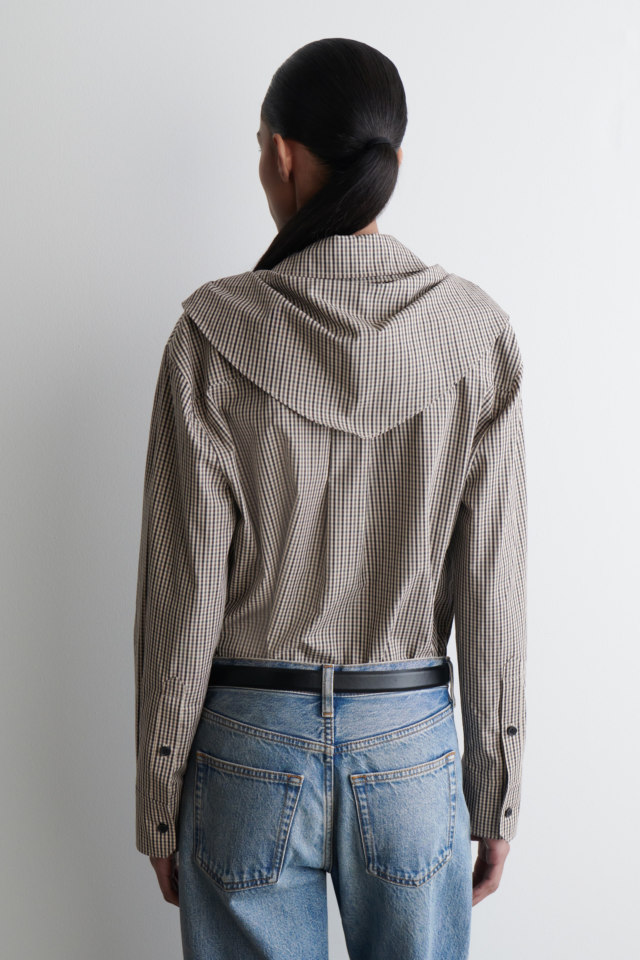 Agrandir l'image: Une chemise micro-gingham marron et crème est vue de dos, avec un détail de col surdimensionné et drapé s'étendant sur les épaules et des manches longues avec des poignets à bouton unique. Elle est portée par-dessus un jean bleu clair avec une ceinture noire.