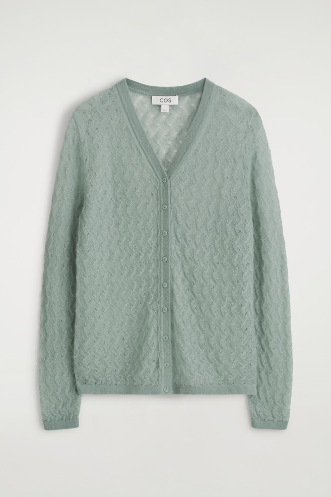 Ein hellgrüner V-Ausschnitt-Cardigan wird von vorne präsentiert. Er zeichnet sich durch ein zartes Pointelle-Strickmuster im gesamten dezent transparenten Stoff aus, mit einer Reihe passender Knöpfe, die die Vorderseite schließen, und gerippten Bündchen und einem gerippten Saum, die das Kleidungsstück abrunden. - 1