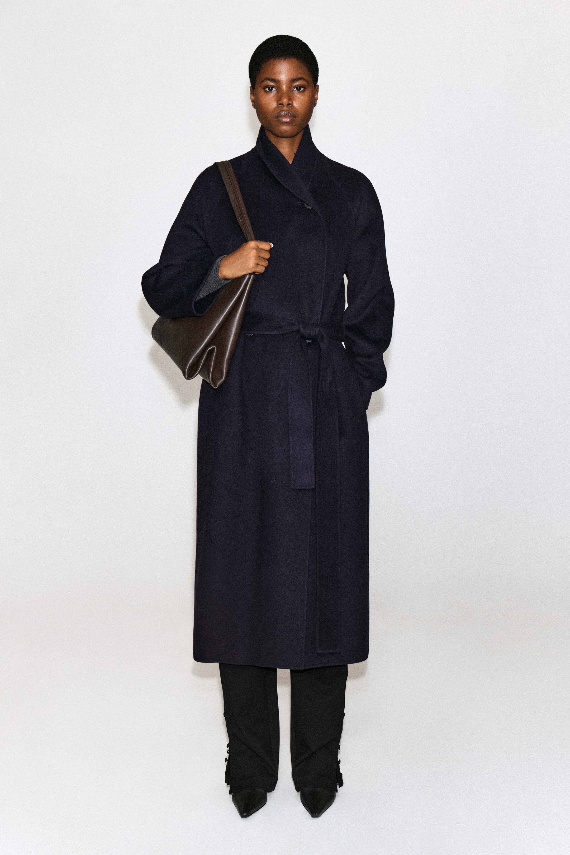 Visualizza l'immagine ingrandita: La modella indossa un cappotto lungo in lana double-face con collo a scialle, in blu navy scuro, chiuso in vita con una cintura annodata nello stesso tessuto. Il cappotto è abbinato a pantaloni neri, scarpe nere con punta affusolata e una borsa a spalla in pelle marrone scuro portata dalla modella.