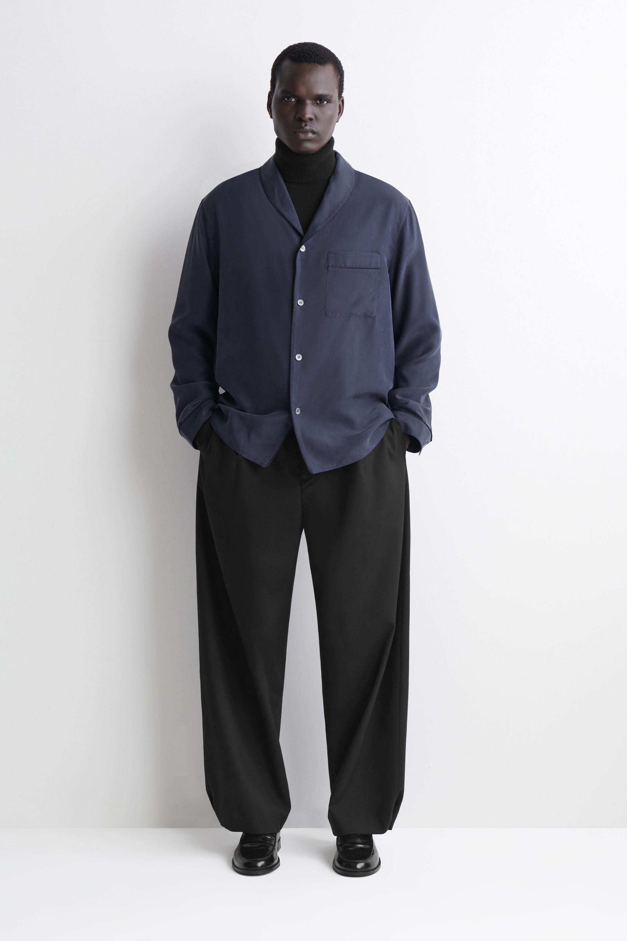 A front-facing, fluid dark-blue silk shirt features a shawl collar, a single chest pocket and light-coloured buttons. The relaxed-fit shirt is layered over a black roll-neck sweater and wide-leg black trousers. A front-facing, fluid dark-blue silk shirt features a shawl collar, a single chest pocket and light-coloured buttons. The relaxed-fit shirt is layered over a black roll-neck sweater and wide-leg black trousers.
