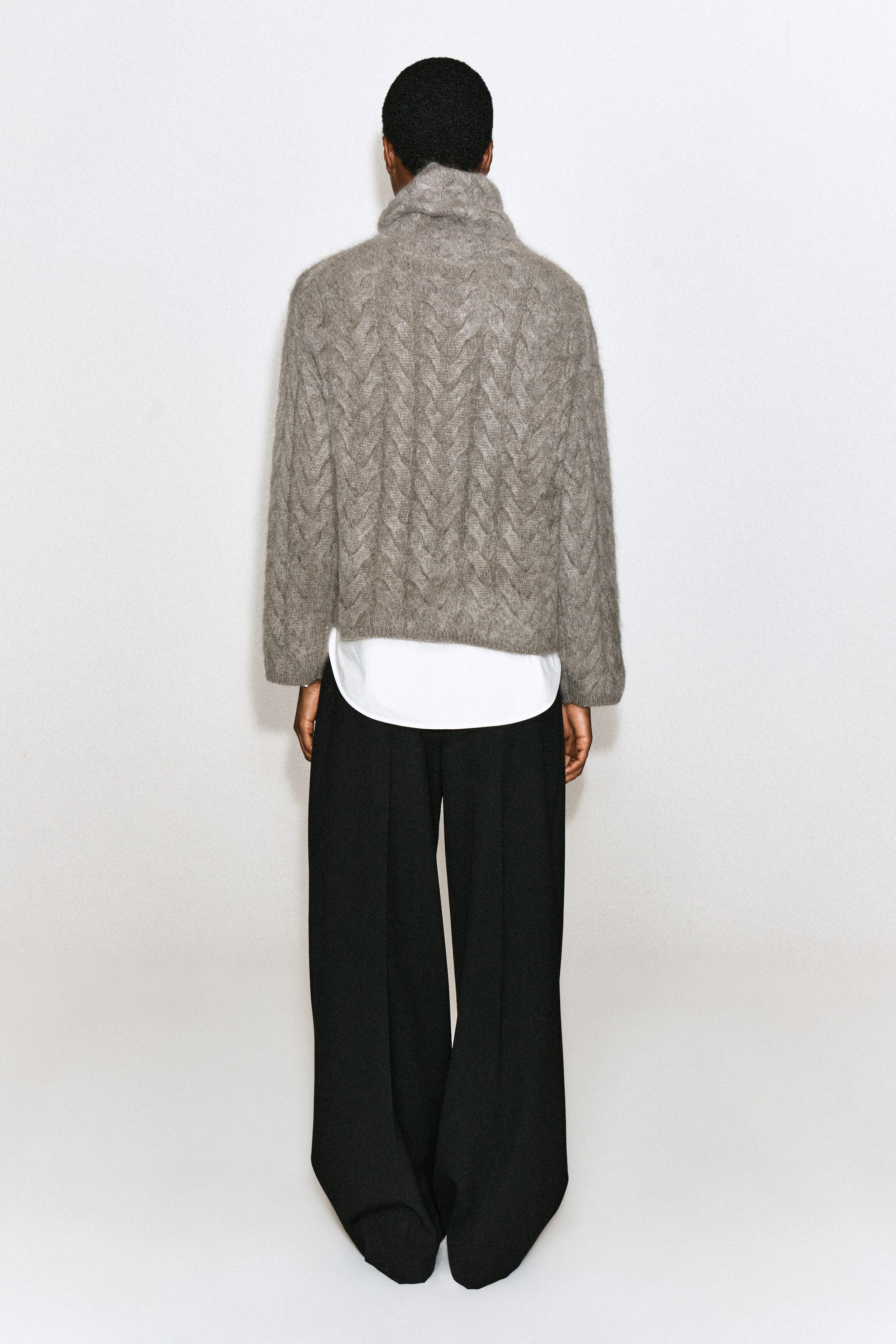 Un chandail à col montant en cachemire brossé, tricoté torsadé, d’une teinte taupe-gris texturée, est porté par le mannequin, vu de dos. Le chandail présente un motif épais de tricot torsadé et un col haut, et il est porté par-dessus une chemise blanche et agencé à un pantalon noir à jambes larges. Un chandail à col montant en cachemire brossé, tricoté torsadé, d’une teinte taupe-gris texturée, est porté par le mannequin, vu de dos. Le chandail présente un motif épais de tricot torsadé et un col haut, et il est porté par-dessus une chemise blanche et agencé à un pantalon noir à jambes larges.