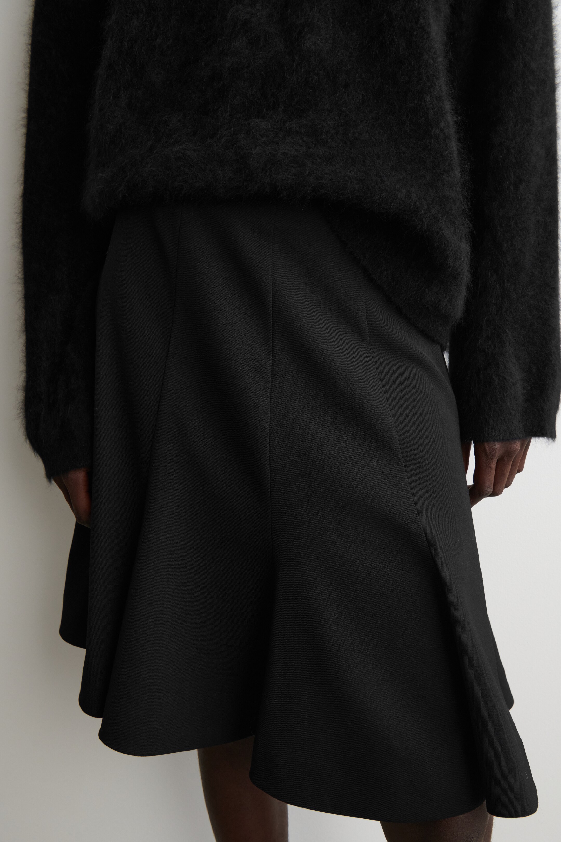 Se større billede: En sort nederdel præsenteres, med et glat stof og lodrette sømme, der skaber en tætsiddende øvre del, der går over i en bred, flæset kant. Den bæres med en sort, fuzzy sweater lagdelt over dens talje.
