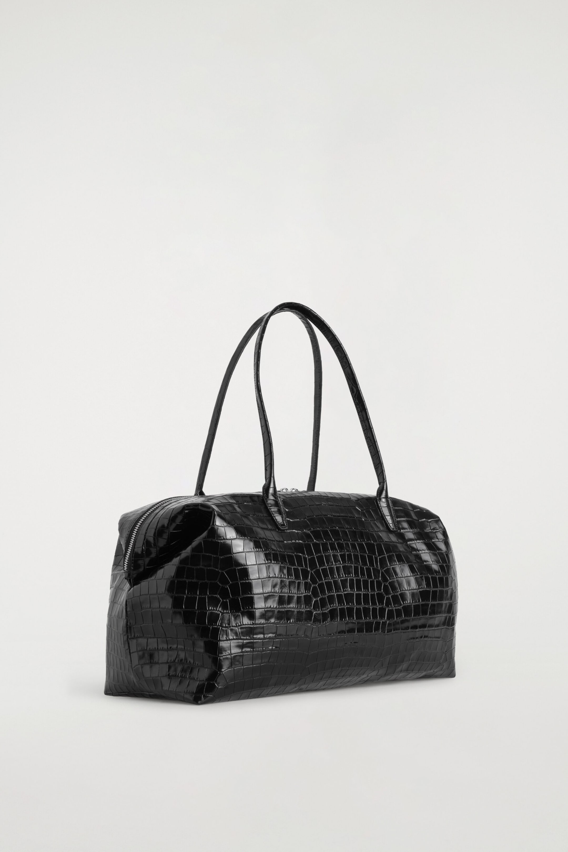 The Frieze Bowling Bag in black croc-effect leather is resting at a three-quarter angle facing front, showcasing its glossy, embossed texture and structured rectangular silhouette. It features two long, slender top handles and a silver-toned zip along the top closure. The Frieze Bowling Bag in black croc-effect leather is resting at a three-quarter angle facing front, showcasing its glossy, embossed texture and structured rectangular silhouette. It features two long, slender top handles and a silver-toned zip along the top closure.