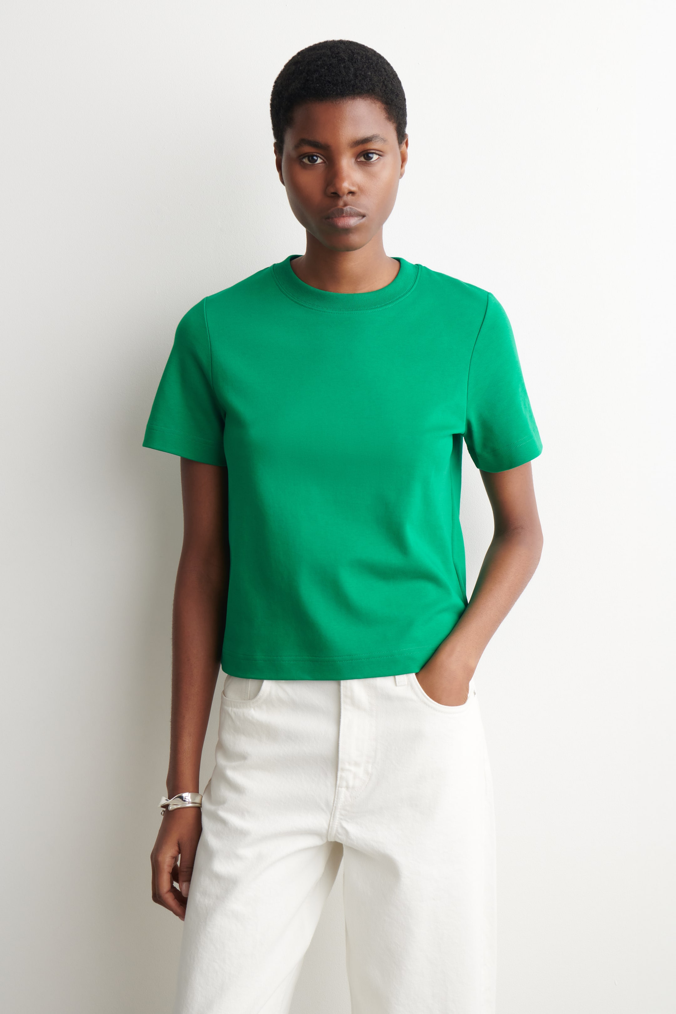 Ingrandisci l'immagine: La t-shirt verde è modellata frontalmente, caratterizzata da un girocollo a coste e maniche corte. Presenta un orlo dritto ed è abbinata a pantaloni bianchi, mettendo in risalto la sua lunghezza corta.