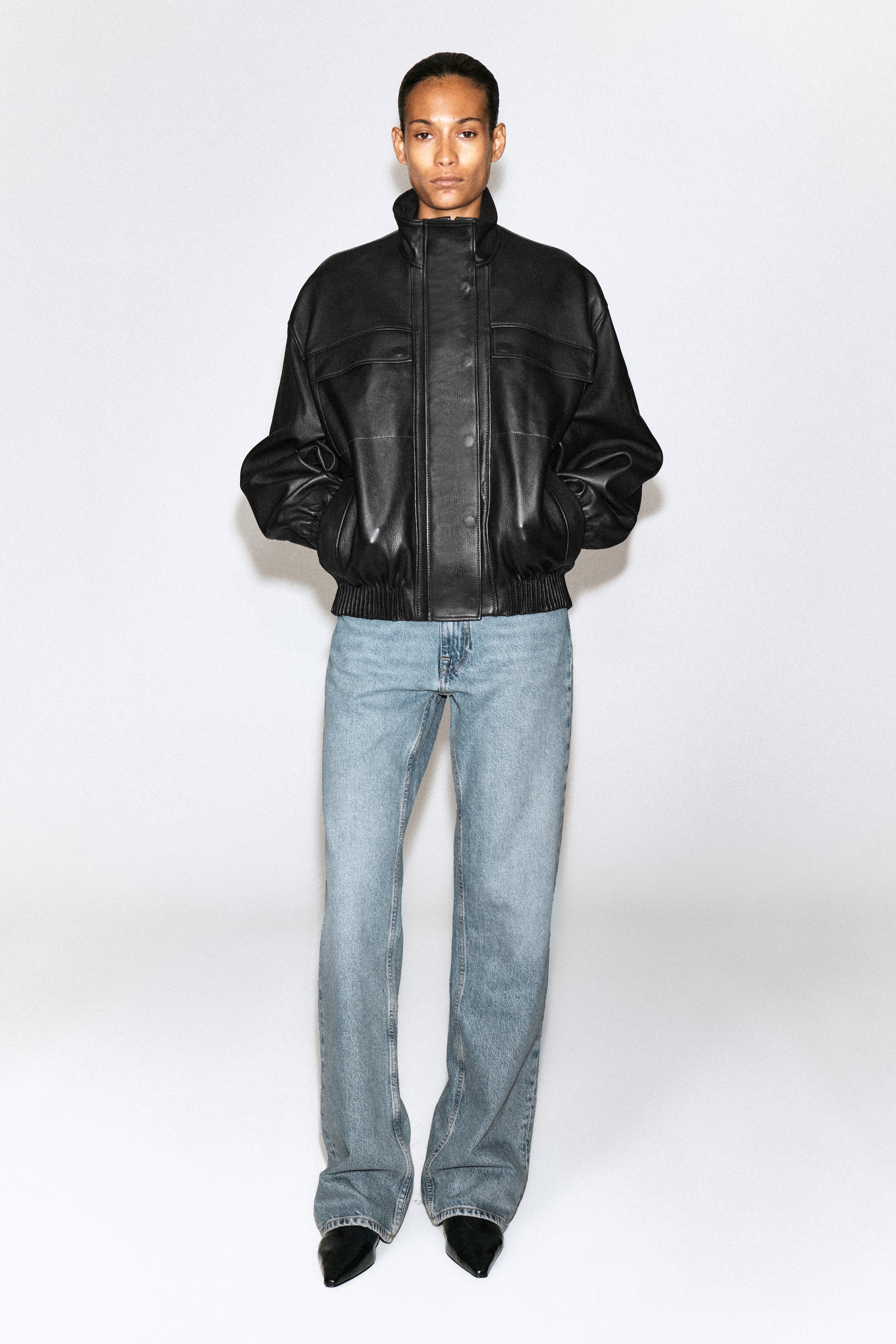 Ver imagem maior: Os jeans bootcut Valley em ganga azul de lavagem clara estão a ser usados pela modelo, apresentando cintura de altura média e perna descontraída que alarga ligeiramente na bainha. Os jeans são combinados com um blusão bomber em pele preto e sapatos pretos de bico fino.