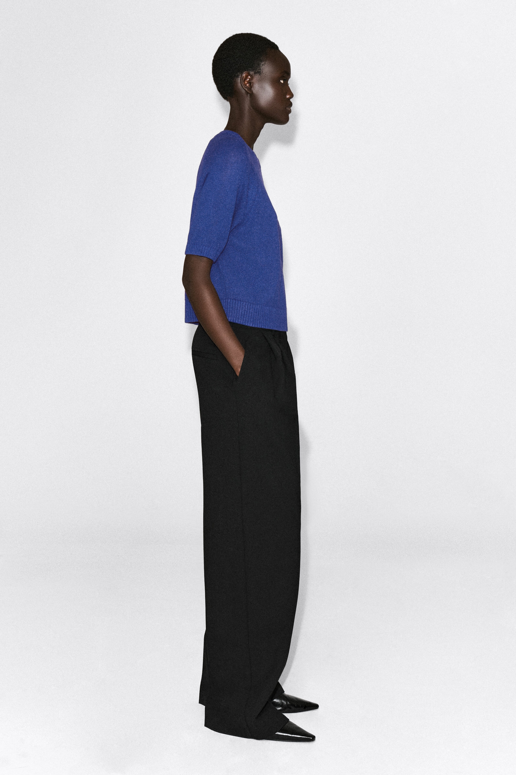 The shrunken knitted linen t-shirt in vibrant blue is shown in side profile facing right, highlighting its cropped fit, crew neck, and short sleeves. The lightweight knit top features a wide ribbed hem that sits just above the waistband of black trousers.