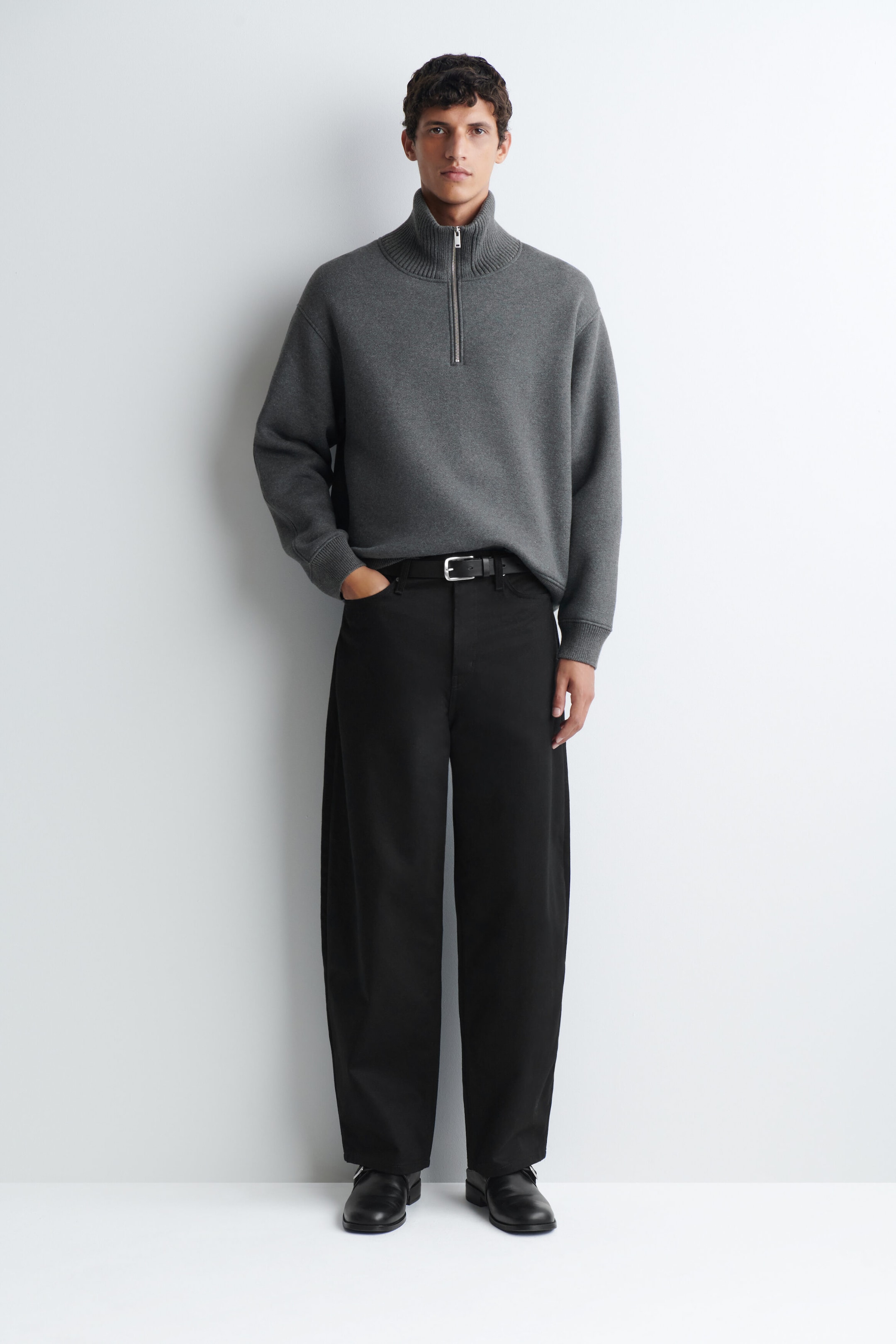 The model is wearing a grey cotton-wool jersey zip-up sweater, featuring a high ribbed collar with a silver half-zip closure and ribbed cuffs. The sweater is styled partially tucked into a pair of black wide-leg trousers with a black leather belt. The model is wearing a grey cotton-wool jersey zip-up sweater, featuring a high ribbed collar with a silver half-zip closure and ribbed cuffs. The sweater is styled partially tucked into a pair of black wide-leg trousers with a black leather belt.