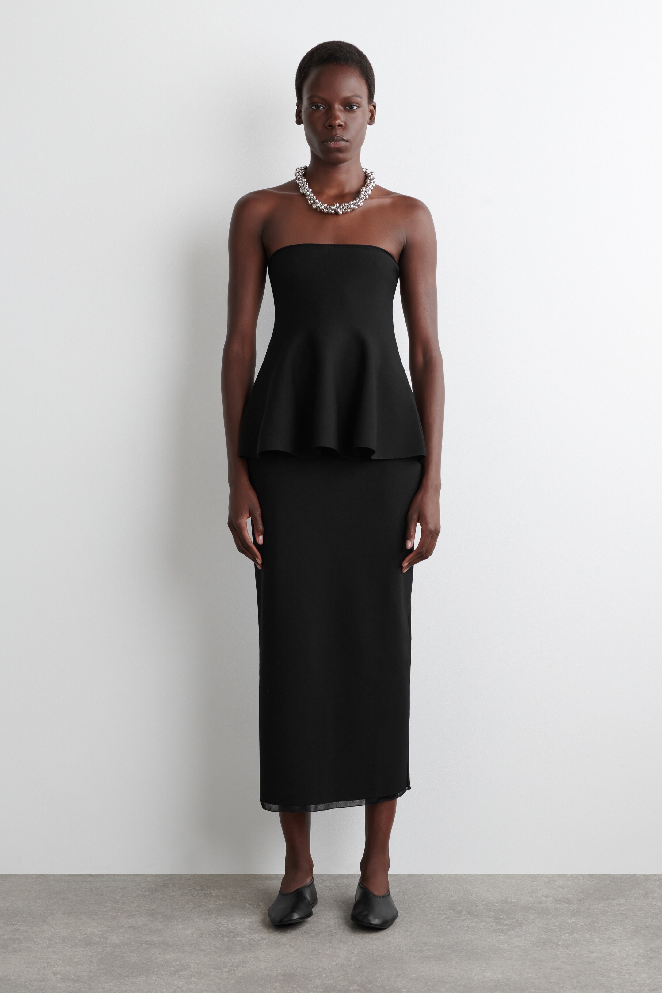 View larger image: A black strapless peplum top, featuring a fitted bodice and a flared peplum hem, is paired with a matching straight-cut midi skirt that includes a subtle sheer hemline, all presented front-facing.