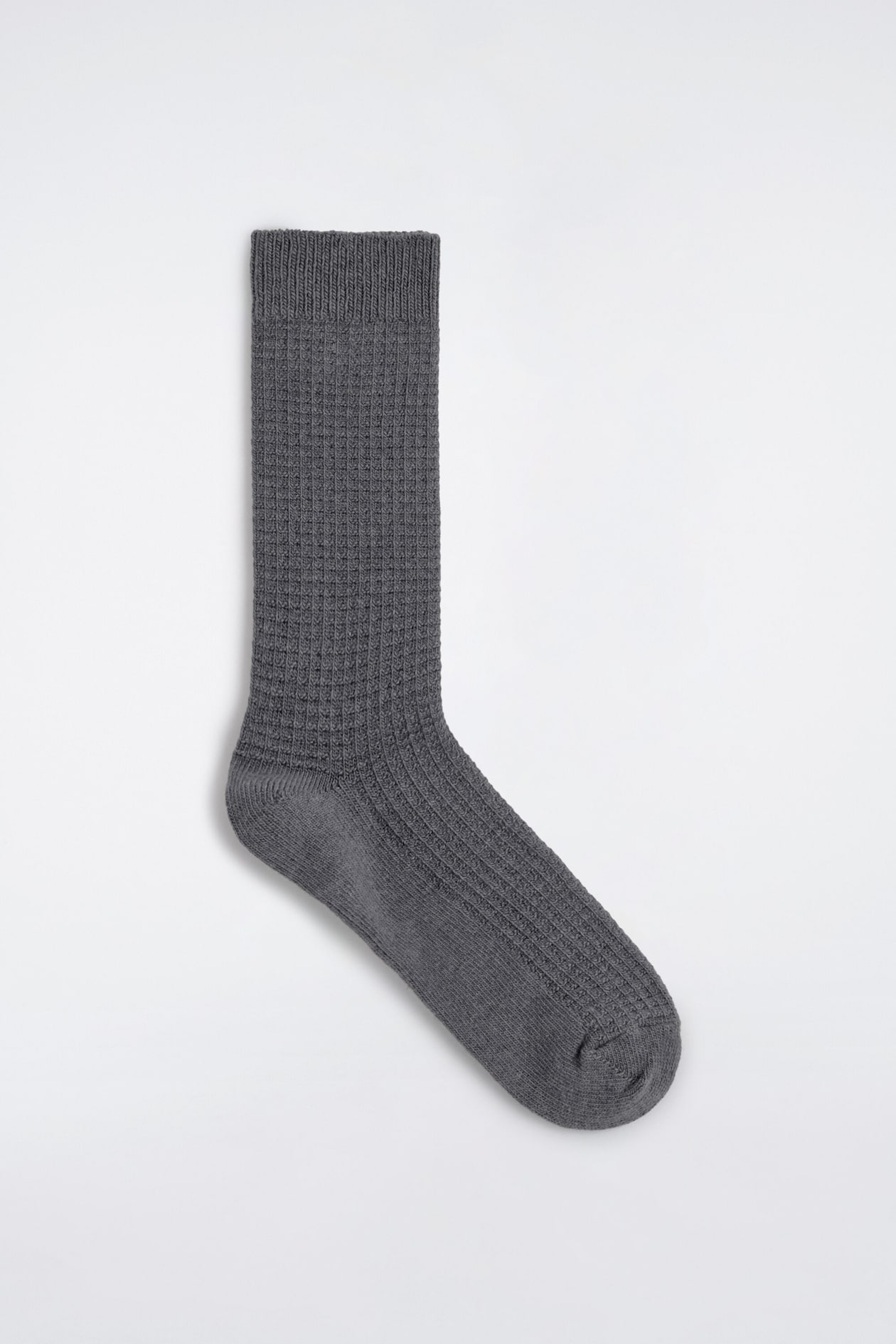 Eine graue Socke liegt flach in seitlicher Profilansicht und zeigt ihre strukturierte Waffelstrick-Optik. Das Design umfasst einen gerippten Bund am oberen Rand, einen Waffelmuster-Schaft und Spann sowie eine glatt gestrickte Ferse und Spitze.