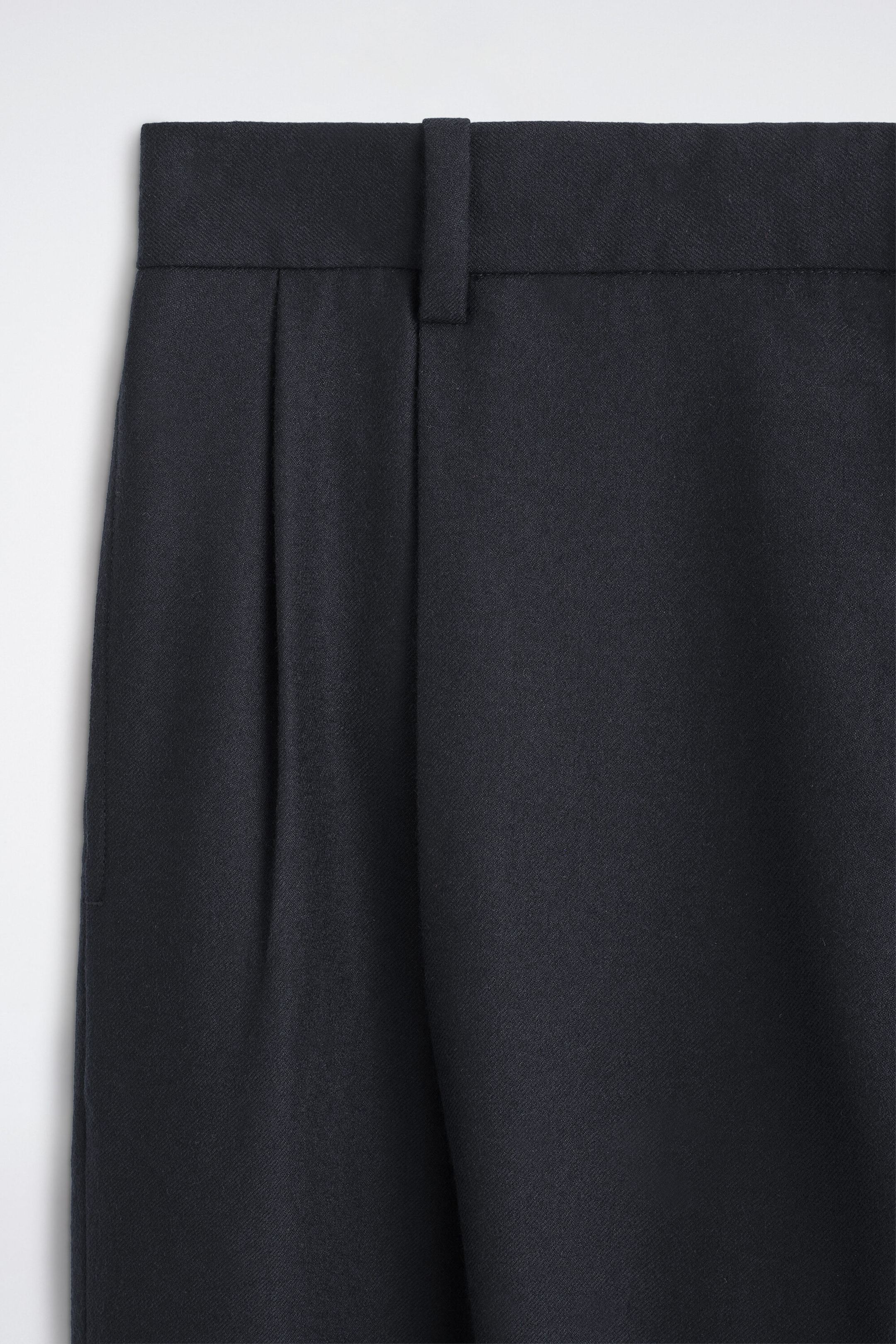Eine Nahaufnahme von vorne zeigt eine dunkelmarineblaue Hose mit einem strukturierten Bund, einer einzelnen Gürtelschlaufe und tiefen Bundfalten, die über den strukturierten Stoff herabfallen. Eine Nahaufnahme von vorne zeigt eine dunkelmarineblaue Hose mit einem strukturierten Bund, einer einzelnen Gürtelschlaufe und tiefen Bundfalten, die über den strukturierten Stoff herabfallen.