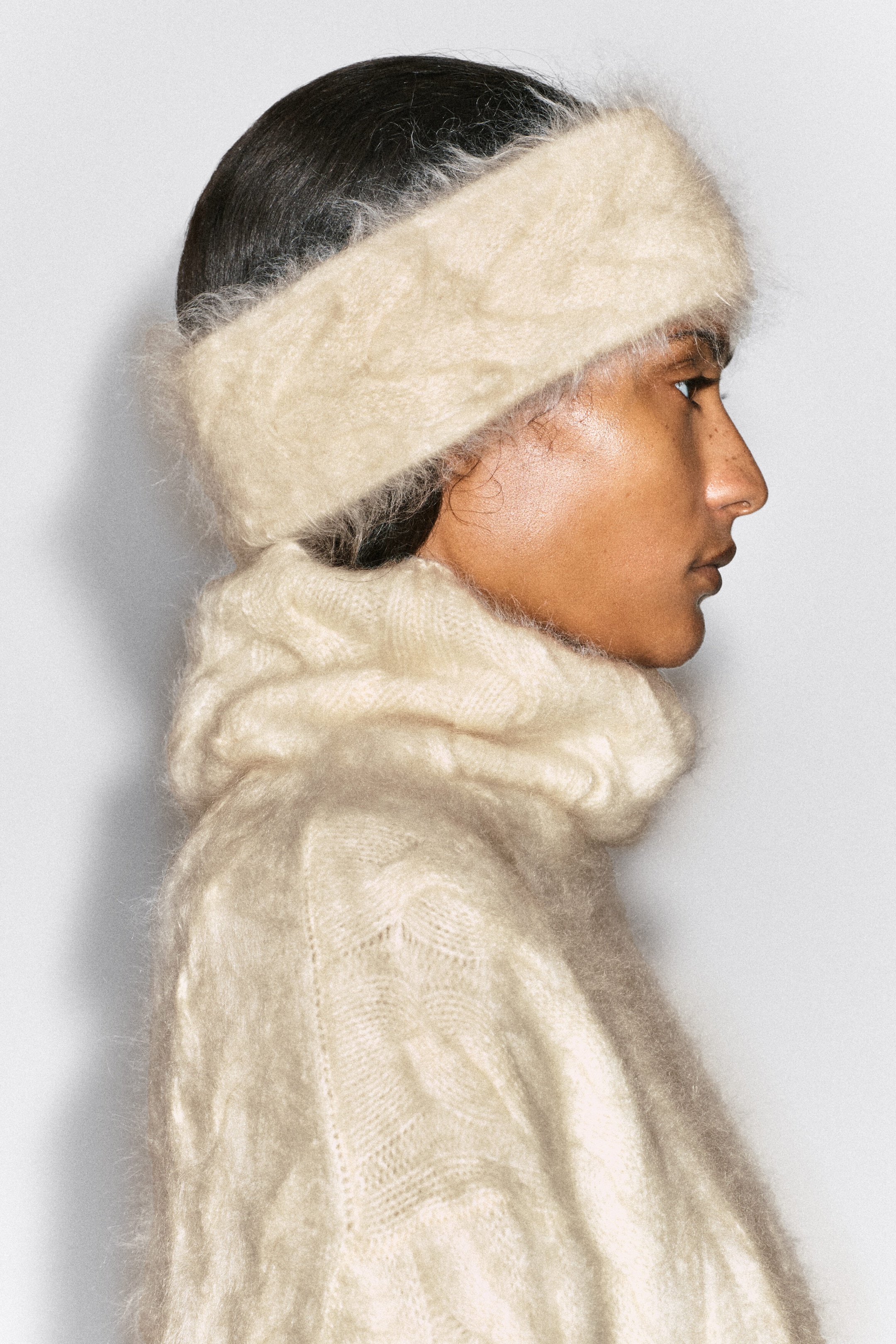 A cream-colored, brushed cable-knit headband is worn by the model over their hair. The headband is paired with a matching high-neck, textured knit sweater in the same off-white shade. A cream-colored, brushed cable-knit headband is worn by the model over their hair. The headband is paired with a matching high-neck, textured knit sweater in the same off-white shade.