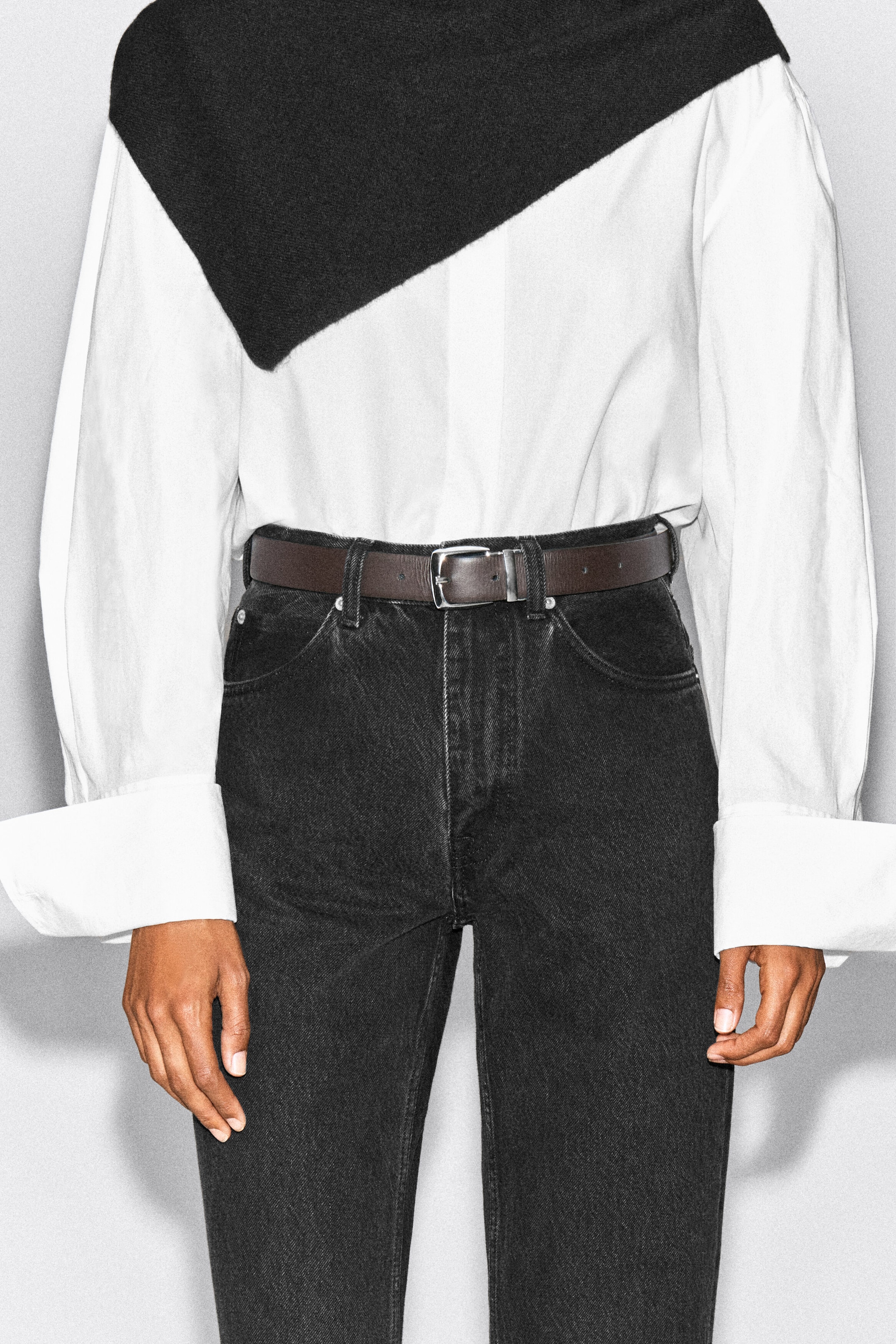 Ein dunkelbrauner Ledergürtel mit einer silberfarbenen rechteckigen Schnalle wird von dem Model durch die Gürtelschlaufen einer schwarzen Jeans getragen. Der Gürtel ist mit einem in die Hose gesteckten weißen Hemd mit Knopfleiste gestylt.