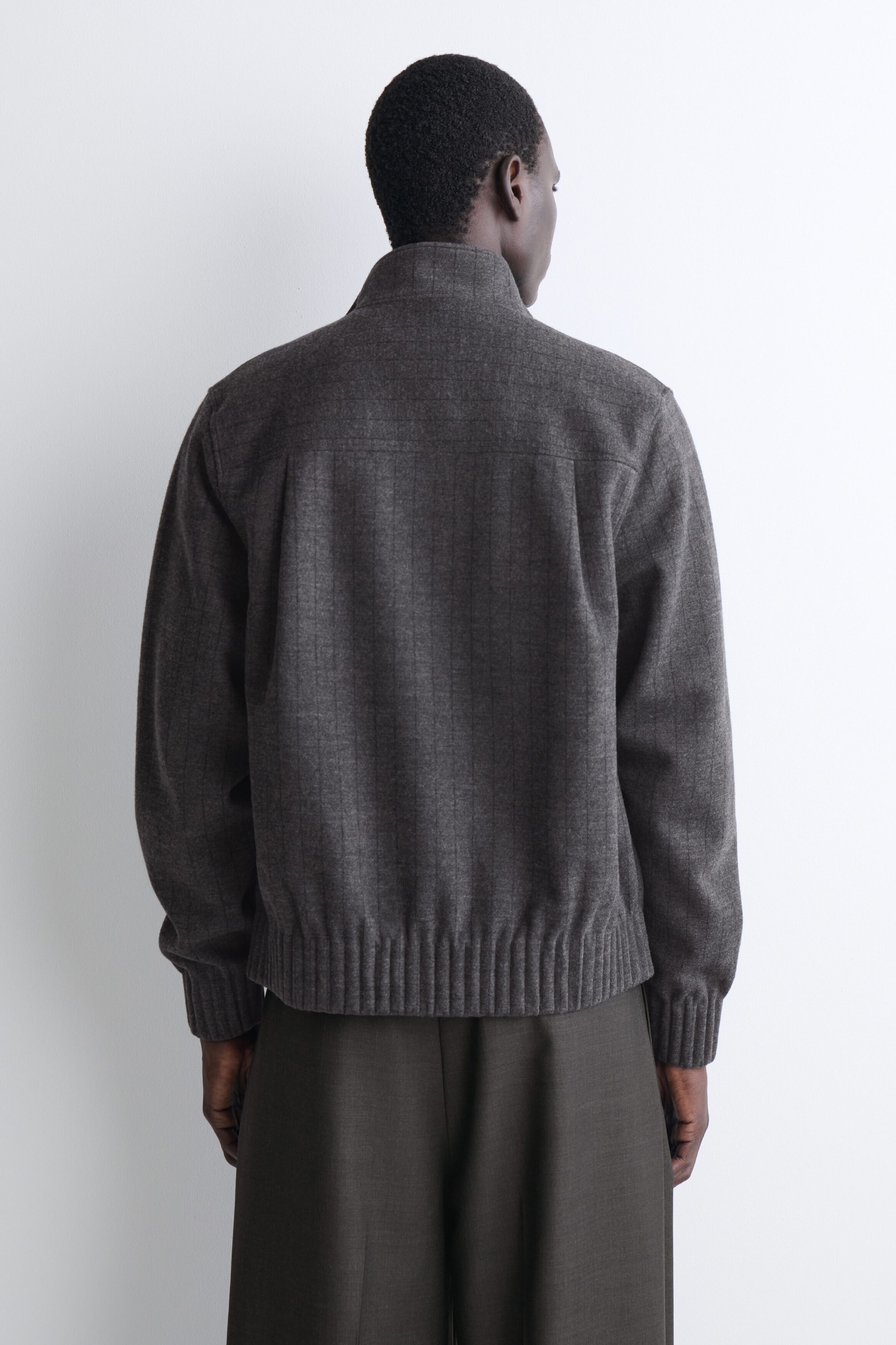 A charcoal gray wool jacket is shown from the back, featuring a high stand-up collar, a horizontal back yoke with subtle pleats, and a faint vertical pinstripe pattern. The jacket has long sleeves with ribbed cuffs and a matching ribbed hem, and is layered over dark, wide-leg trousers. A charcoal gray wool jacket is shown from the back, featuring a high stand-up collar, a horizontal back yoke with subtle pleats, and a faint vertical pinstripe pattern. The jacket has long sleeves with ribbed cuffs and a matching ribbed hem, and is layered over dark, wide-leg trousers.