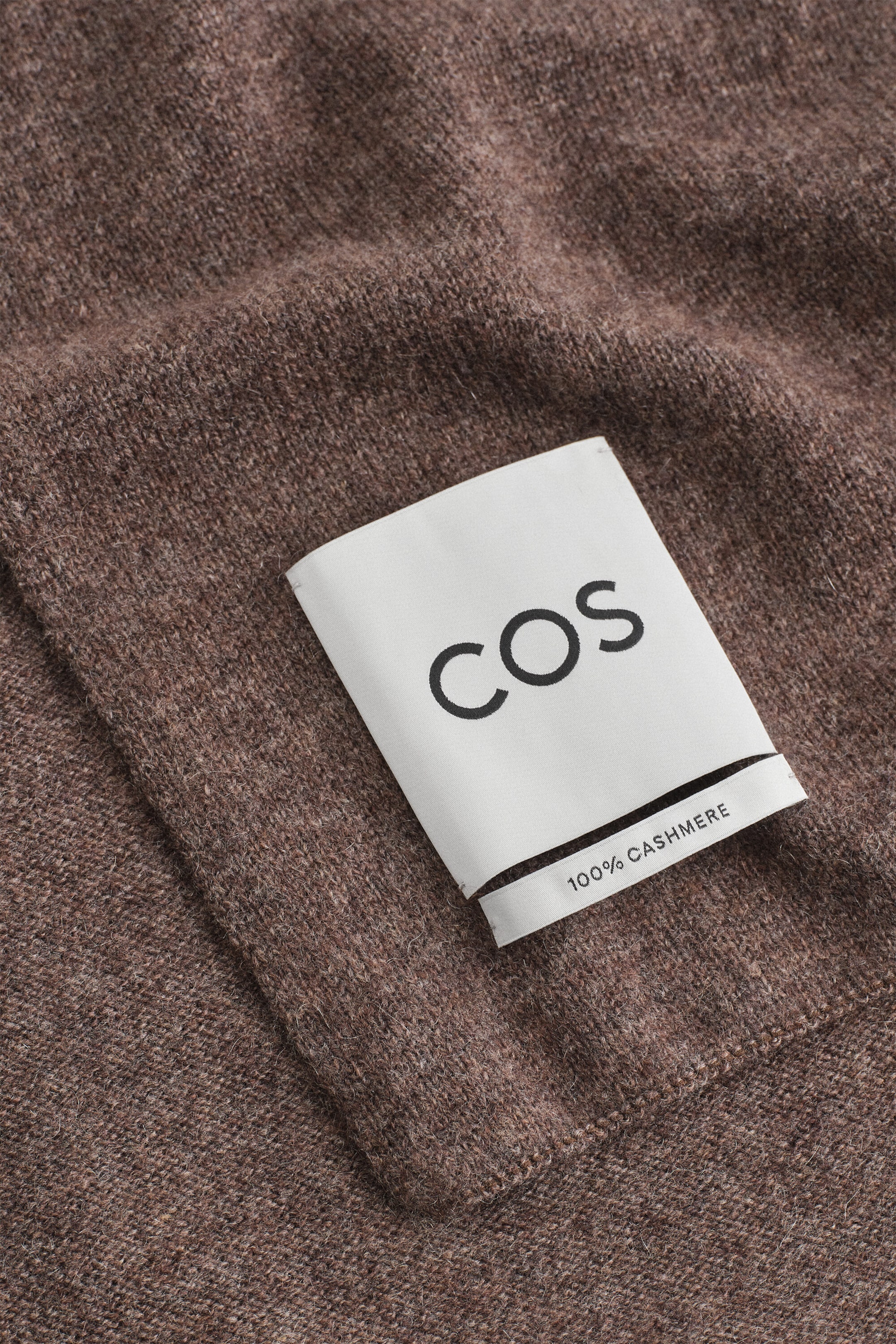 A cashmere scarf in heathered brown showcases its soft, finely knit texture in a close-up view. A white fabric label reading "COS" in black letters and a smaller tag reading "100% CASHMERE" are stitched onto the fabric. A cashmere scarf in heathered brown showcases its soft, finely knit texture in a close-up view. A white fabric label reading "COS" in black letters and a smaller tag reading "100% CASHMERE" are stitched onto the fabric.