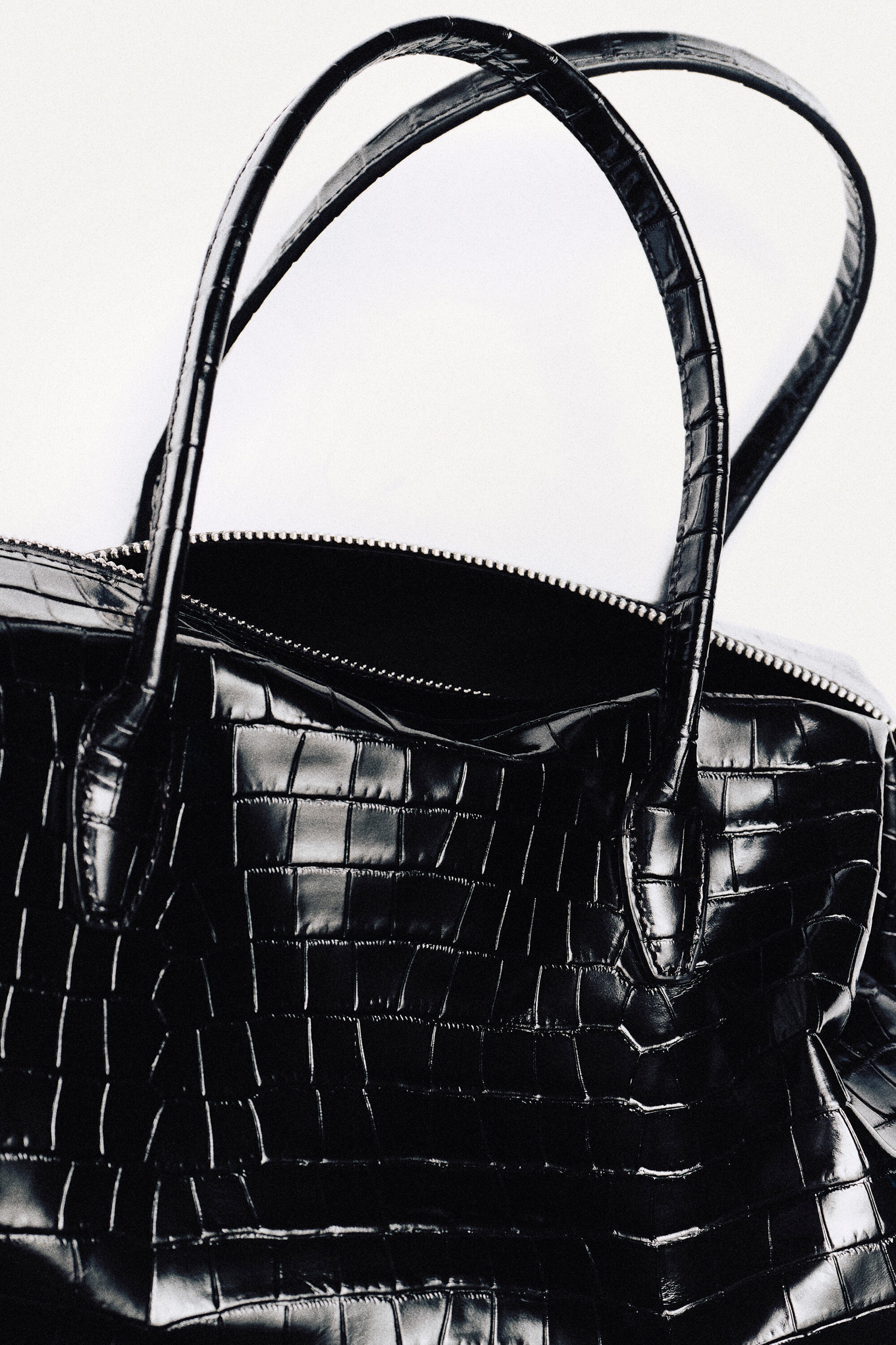 The Montmartre bowling bag in black croc-effect leather features a glossy, embossed crocodile texture and dual arched top handles in a close-up front view. A silver-toned zipper runs along the top opening, partially revealing the dark interior.