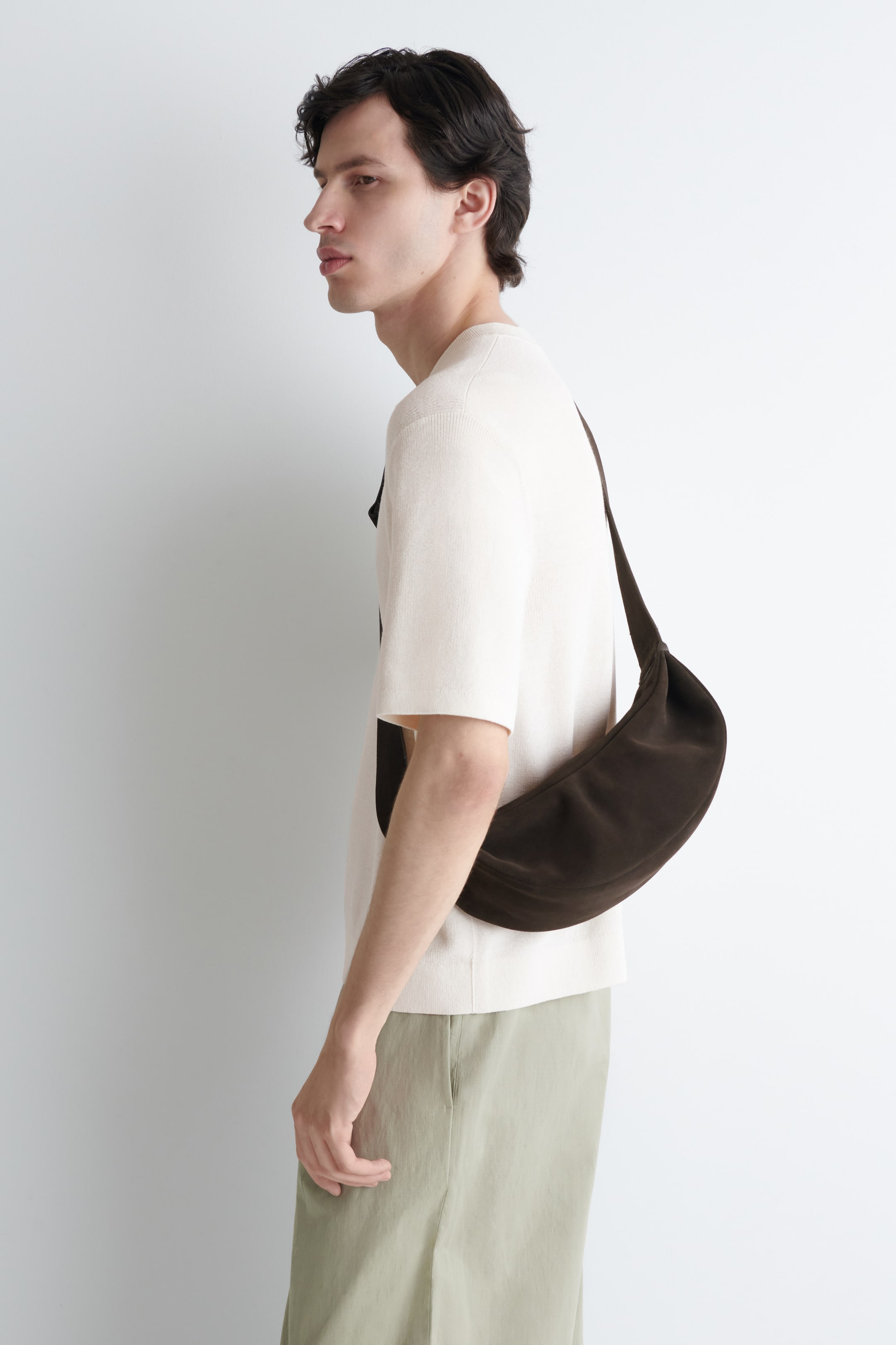 Visualizza immagine più grande: Una borsa a tracolla a forma di mezzaluna di colore marrone scuro viene indossata trasversalmente sul corpo, con la sua forma morbida e non strutturata che si adagia sul fianco.