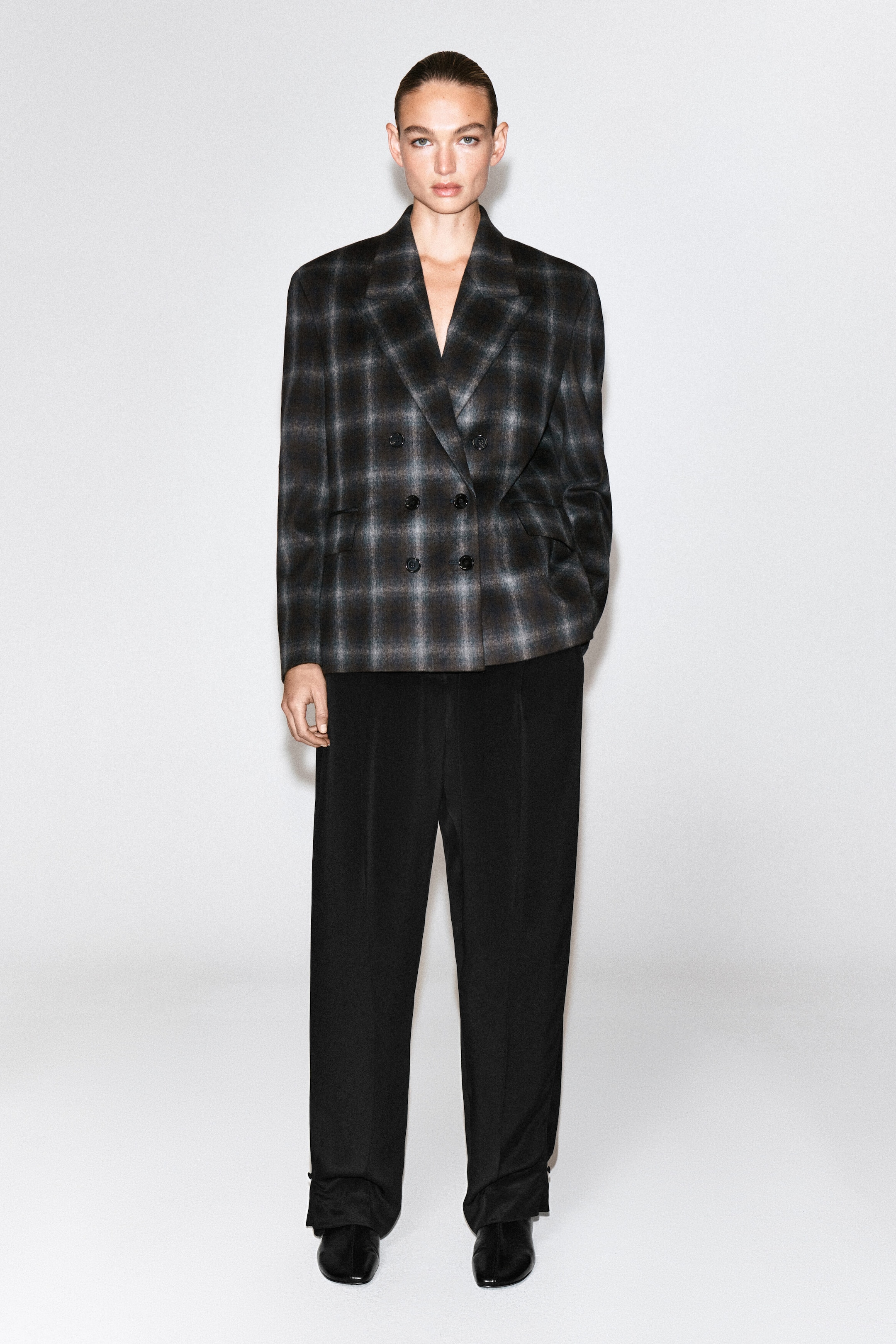 Un blazer croisé en mélange de laine à motif écossais est porté par le mannequin. Le blazer est doté de revers crantés, d’une fermeture croisée à boutons et de poches latérales, et il est associé à un pantalon noir à jambes larges.