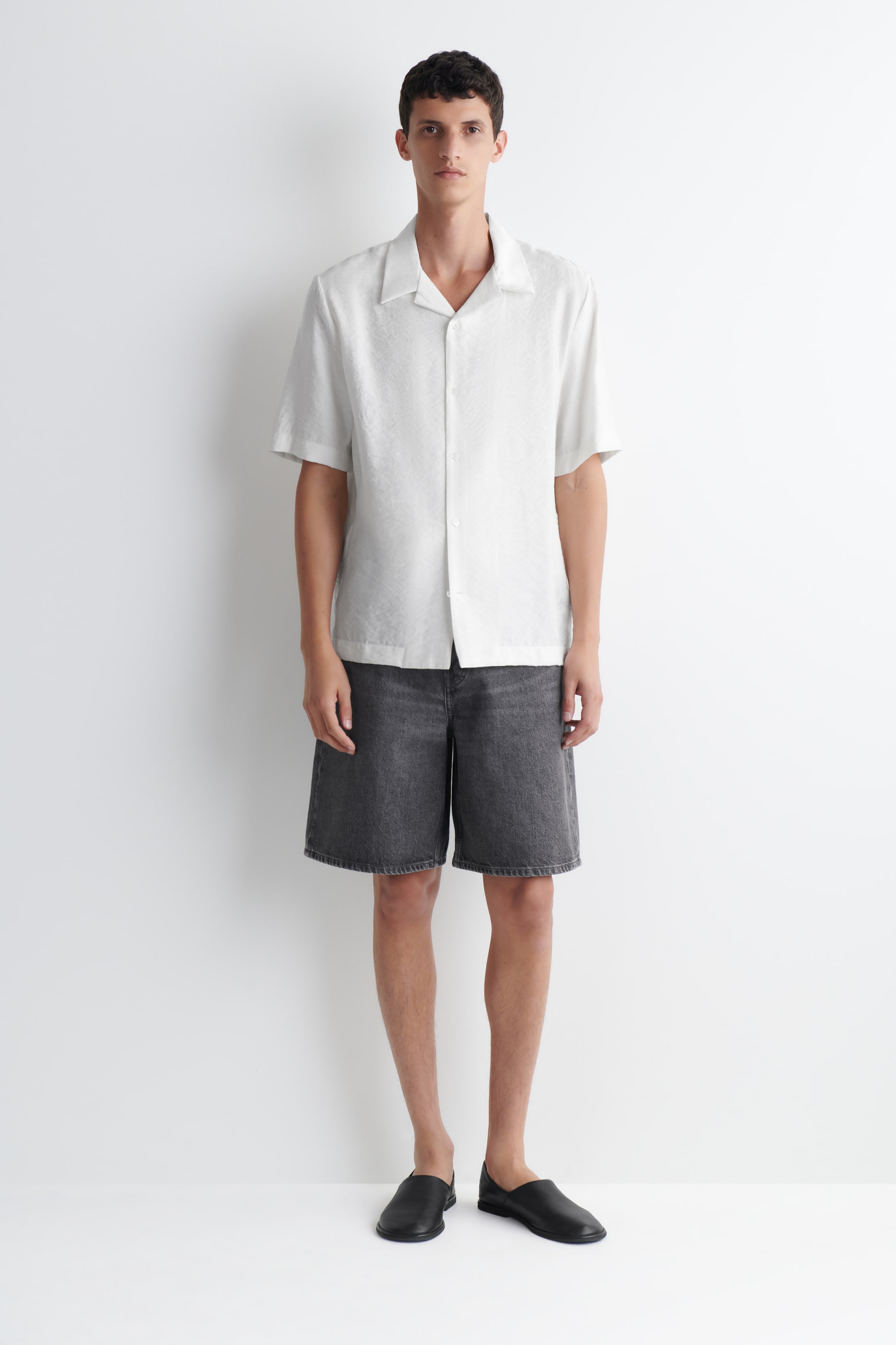 Ver imagen más grande: Los shorts de ciclista en denim negro deslavado están siendo llevados por la modelo. Los shorts relajados de pierna recta llegan por encima de la rodilla, presentan un sutil pespunteado y están combinados con una camisa blanca de manga corta.