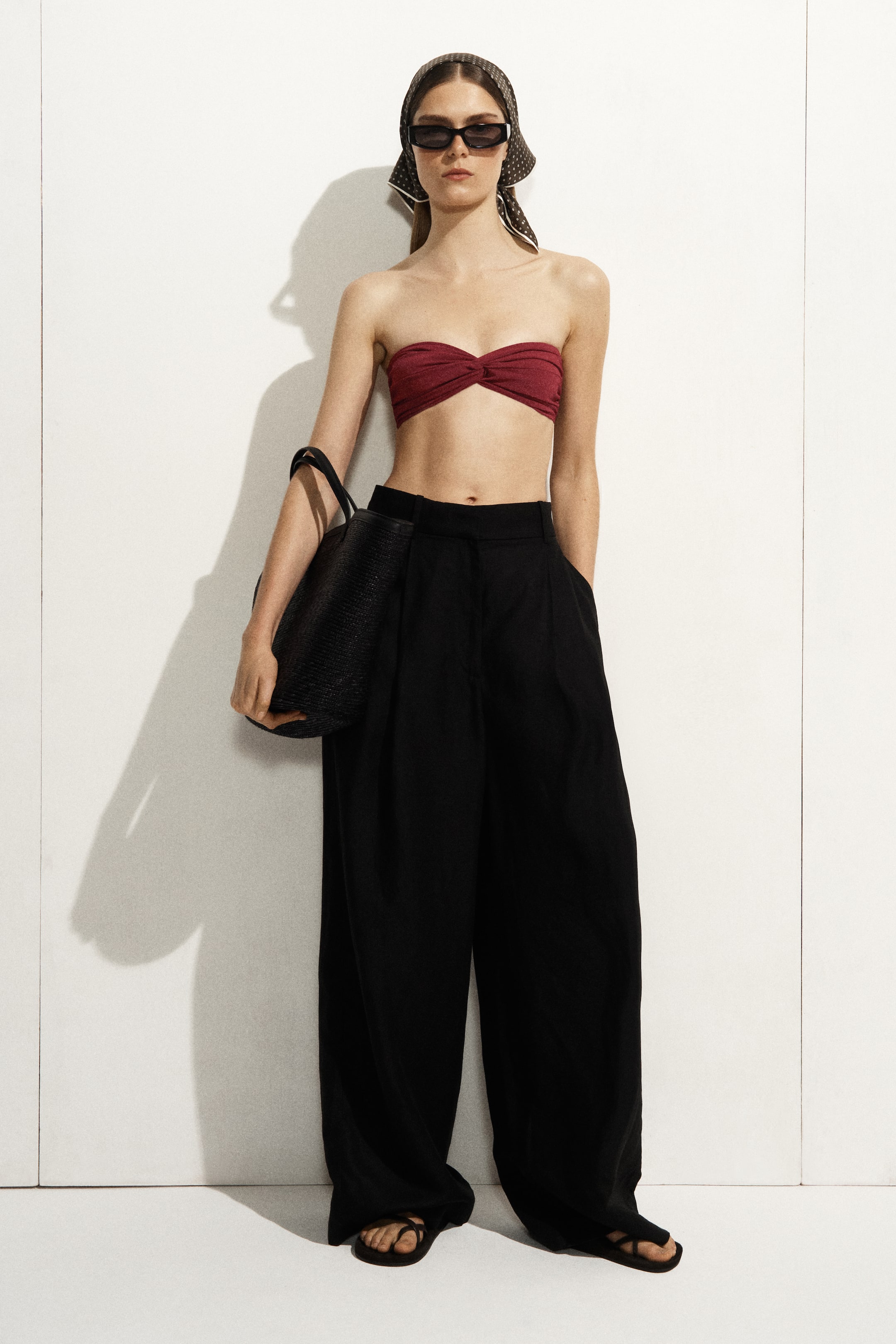 Ver imagen más grande: Se presenta un conjunto de cuerpo entero de frente, que incluye un top bandeau sin tirantes de color rojo intenso con un detalle retorcido en el busto, pantalones de pierna ancha de talle alto en negro, un bolso tote tejido en negro, sandalias planas negras, un pañuelo estampado y gafas de sol rectangulares.