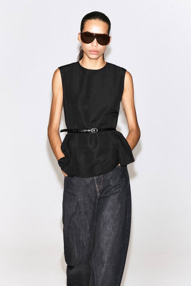 A black sleeveless pleated peplum top is shown from the front, featuring a round neck and structured shoulders. The top is styled with a thin black belt cinched at the waist to accentuate the flared, pleated peplum hem. - 1