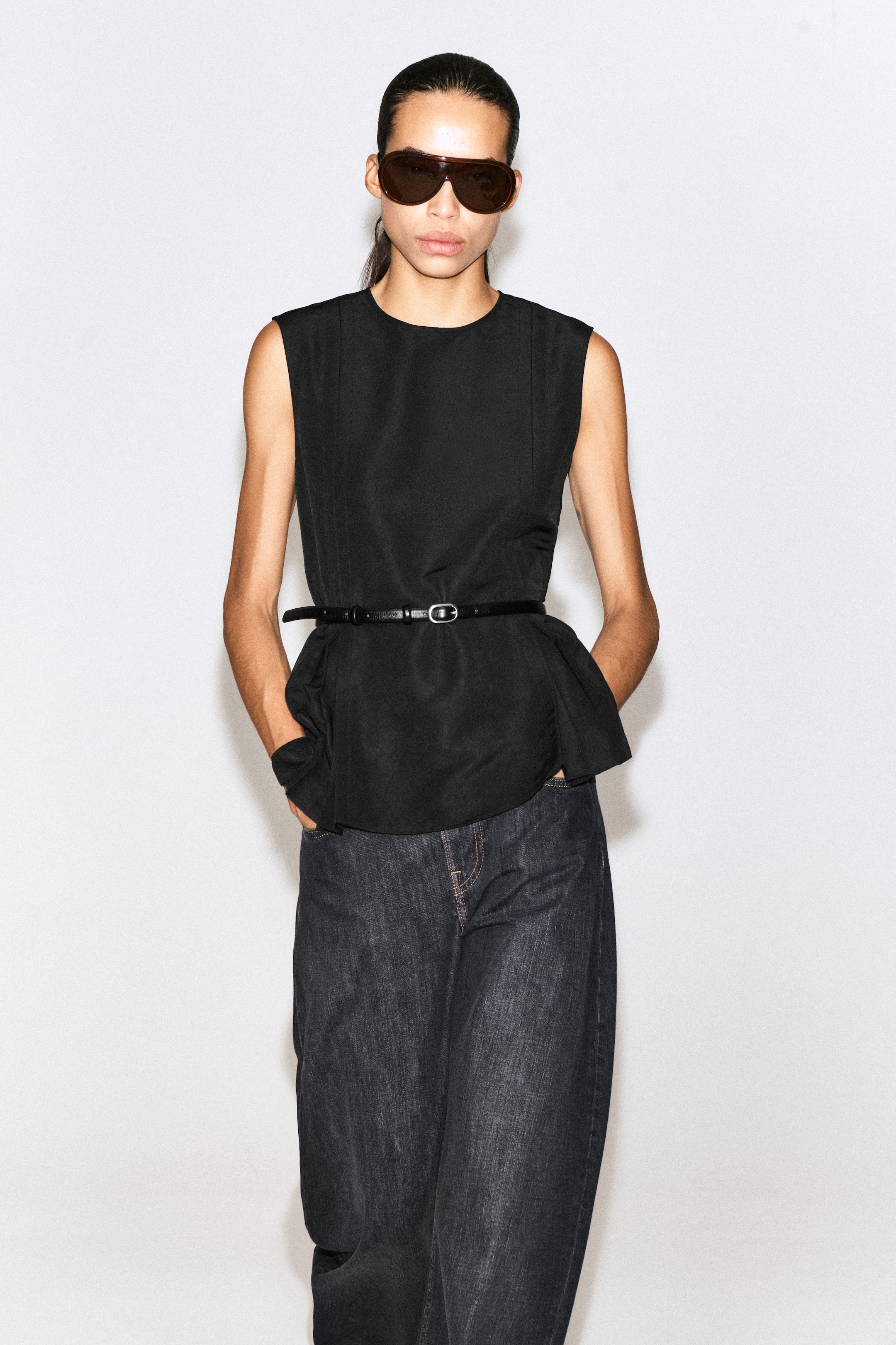 Visualizza l'immagine ingrandita: Un top peplum nero senza maniche viene mostrato frontalmente, con collo rotondo e spalle strutturate. Il top è abbinato a una sottile cintura nera stretta in vita per accentuare l’orlo peplum svasato e plissettato.