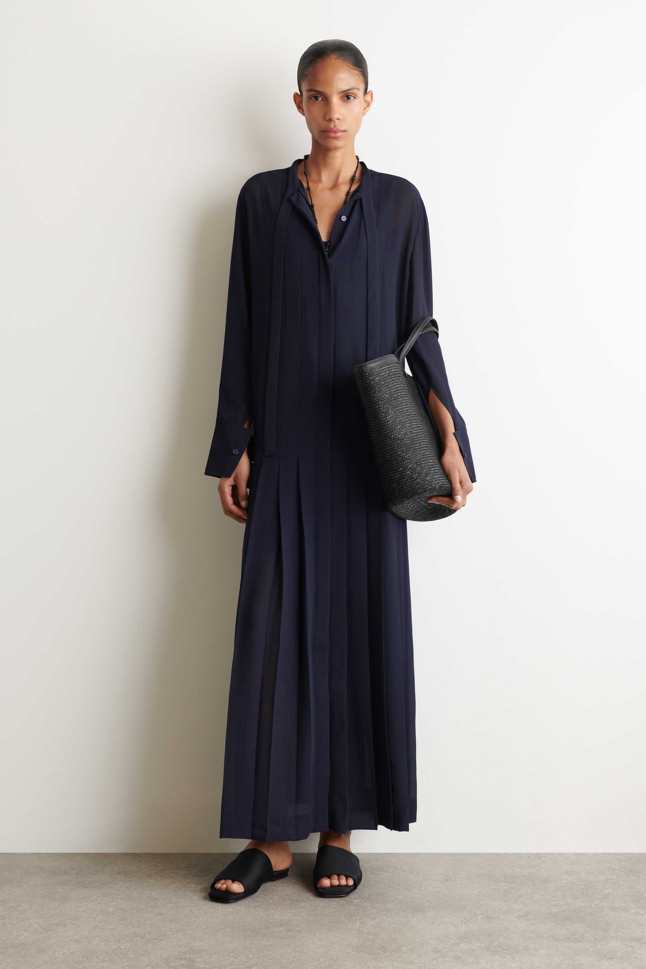 Agrandir l'image: Une robe maxi bleu marine présente un col montant avec une patte de boutonnage ouverte et des manches longues avec des poignets boutonnés et des fentes aux poignets. Des plis nervurés verticaux ornent le corsage, tandis que la jupe fluide met en valeur des plis plats.