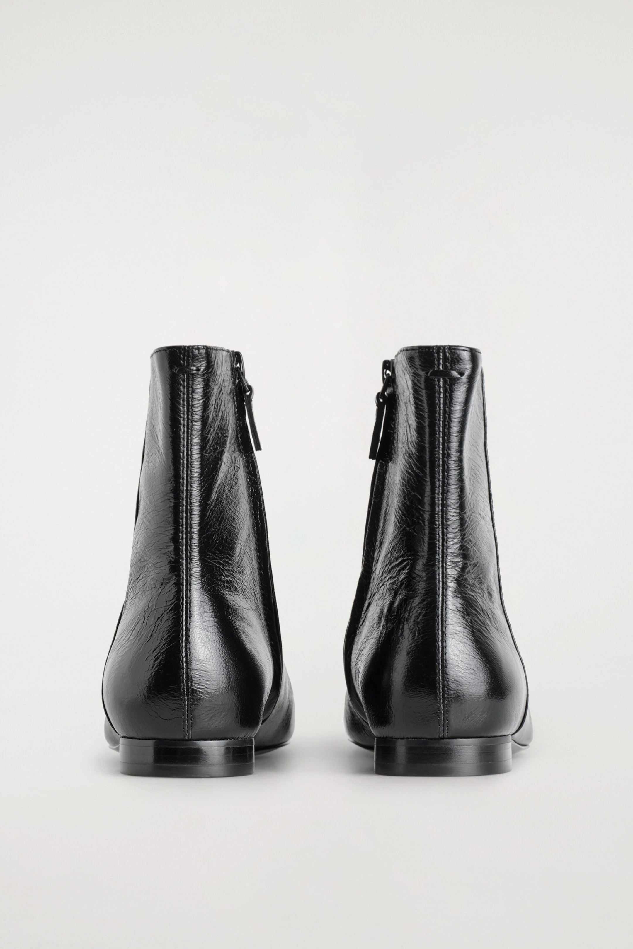 Black ankle boots are facing backwards, showcasing a glossy, crinkled leather finish and low, flat heels. They display a vertical seam running down the centre back and subtle side zip fastenings.