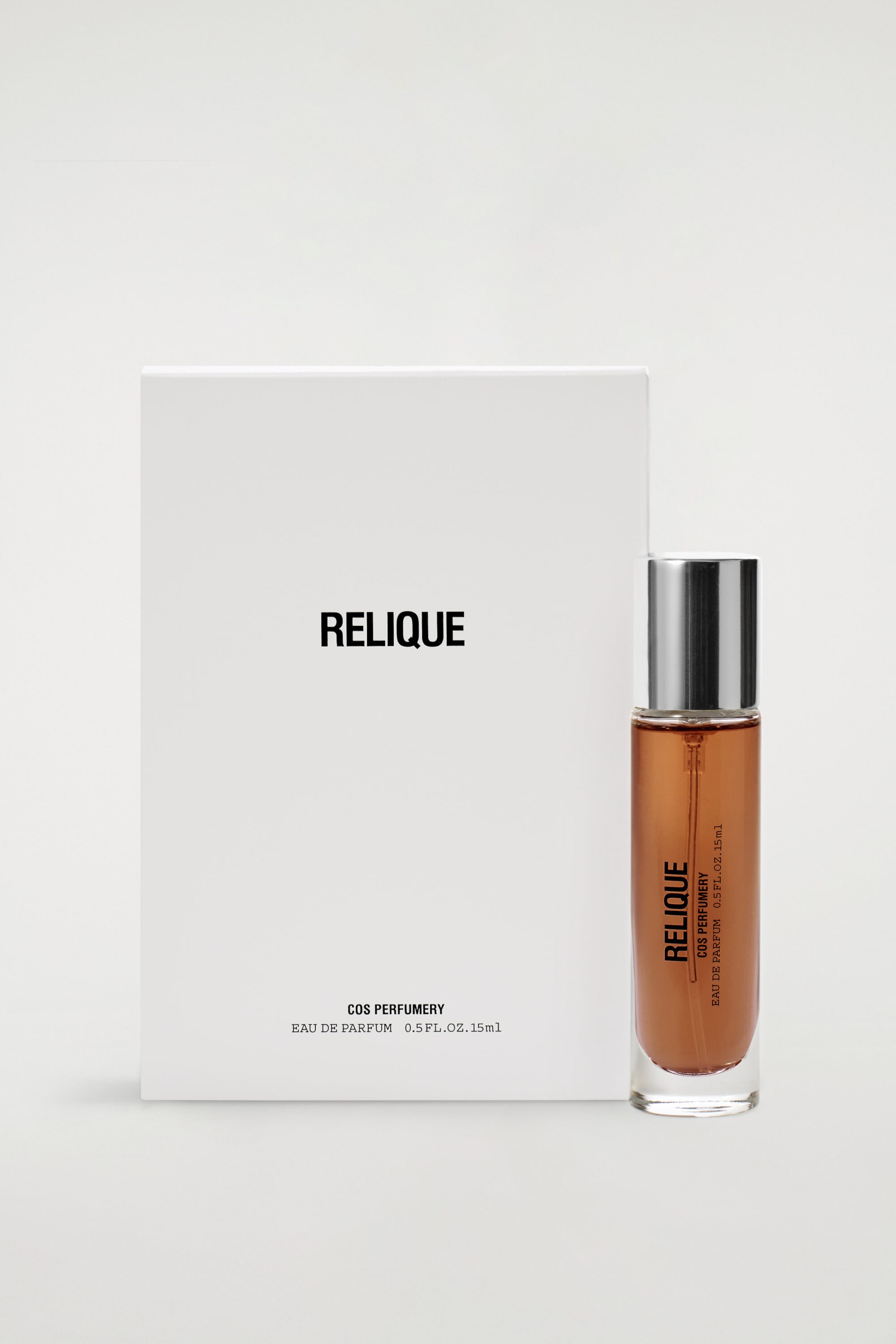 Debout, face avant, l’Eau de Parfum Relique COS Perfumery, 15 ml, est présentée dans un flacon en verre cylindrique et élancé contenant un liquide ambré, surmonté d’un capuchon métallique argenté. Une boîte blanche rectangulaire au design minimaliste est placée à côté du flacon, affichant une identité visuelle noire en sans-serif assortie ainsi que les spécifications du produit.
