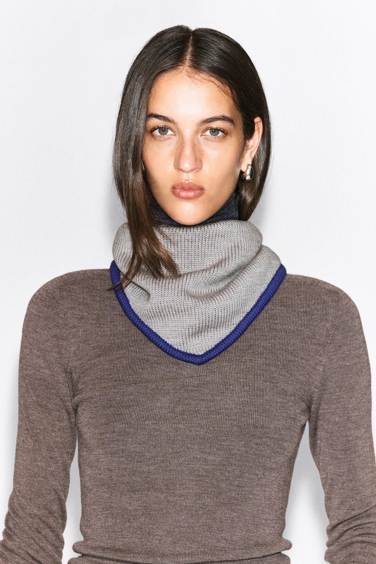 Un paÃ±uelo triangular de lana merina en gris claro con borde azul y remates en contraste se lleva alrededor del cuello de la modelo. El paÃ±uelo de punto canalÃ© cae hasta formar una punta en el pecho, superpuesto sobre un top de punto marrÃ³n de manga larga. - 1