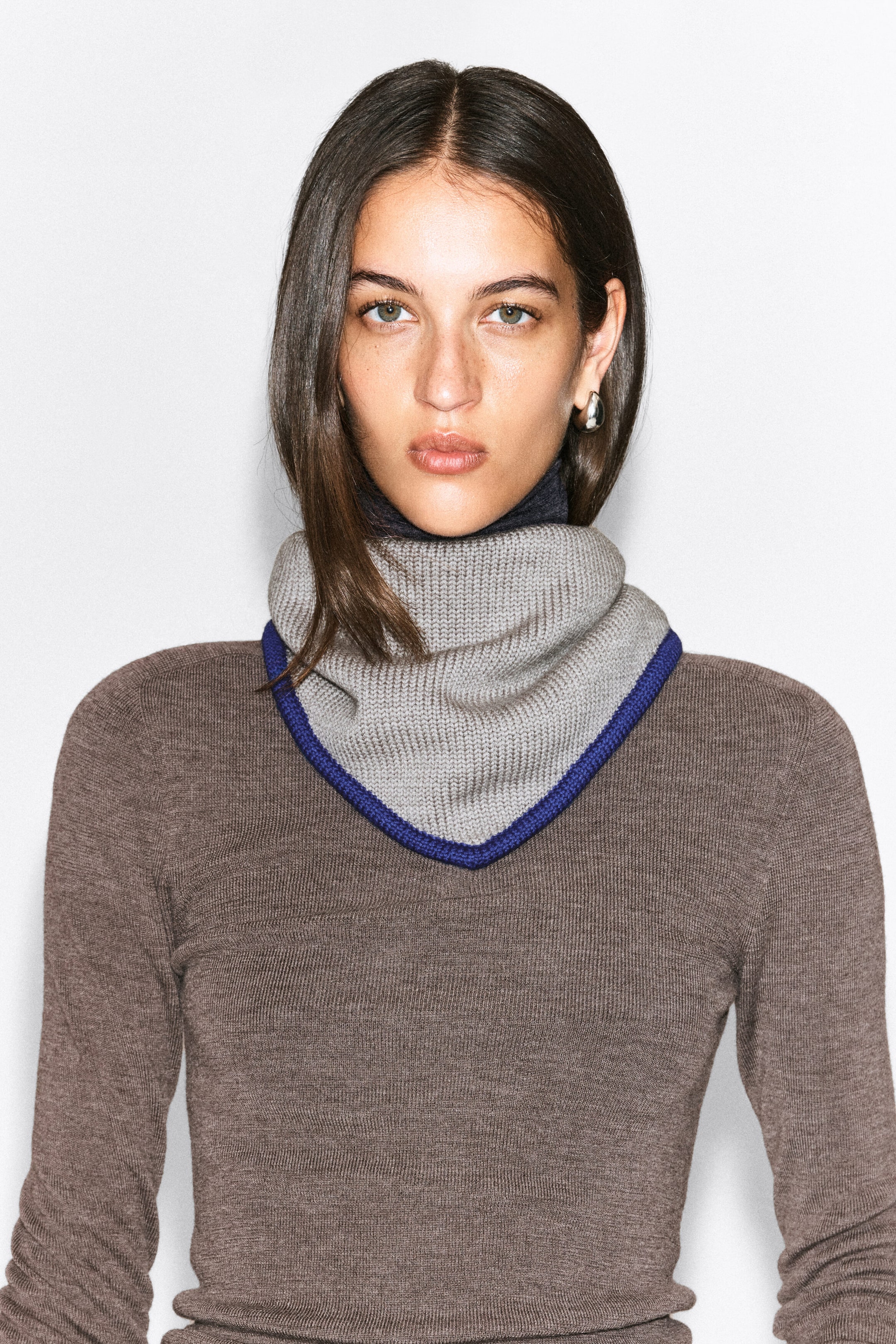A contrast-tipped merino wool triangle scarf in light grey with a blue border is being worn around the neck by the model. The ribbed knit scarf drapes down to a point at the chest, layered over a brown long-sleeved knit top. A contrast-tipped merino wool triangle scarf in light grey with a blue border is being worn around the neck by the model. The ribbed knit scarf drapes down to a point at the chest, layered over a brown long-sleeved knit top.