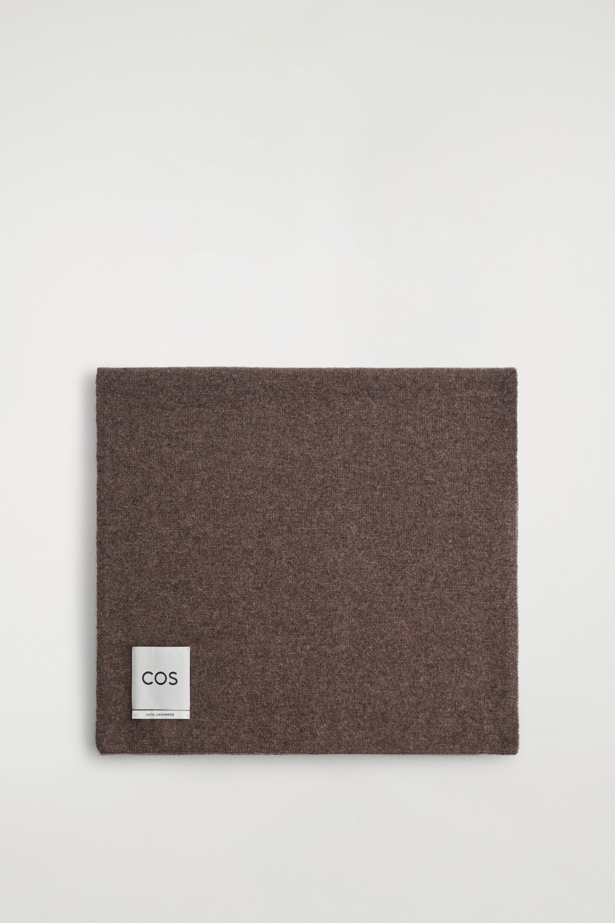 A brown cashmere scarf is laid flat and folded into a square shape, facing forward to show its fine knit texture. A small white rectangular label is stitched near the bottom-left corner.