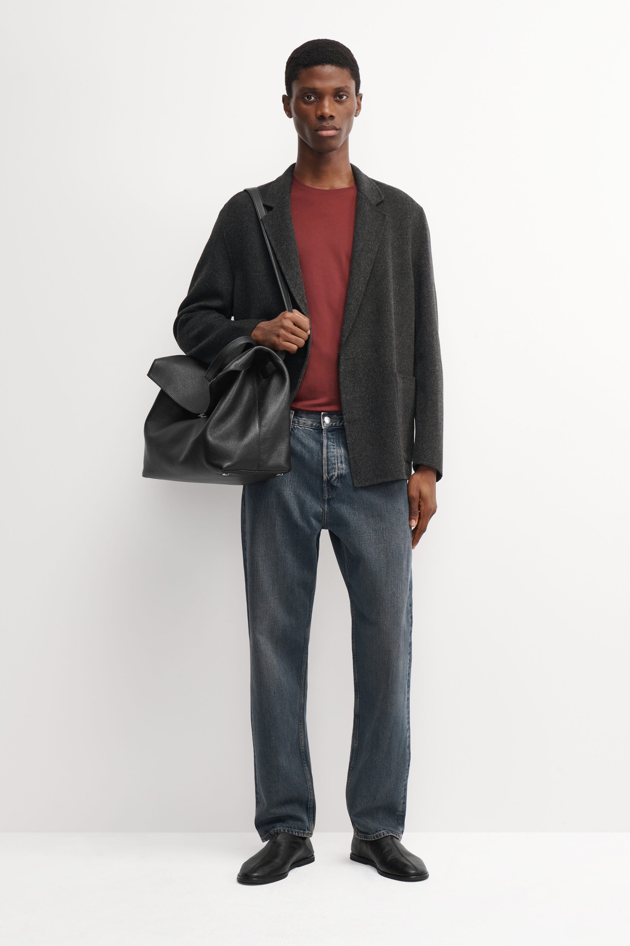 The model is wearing a reddish-brown cotton crew-neck T-shirt tucked into blue jeans and layered under a dark gray casual blazer. They are carrying a black leather shoulder bag while showcasing the outfit. The model is wearing a reddish-brown cotton crew-neck T-shirt tucked into blue jeans and layered under a dark gray casual blazer. They are carrying a black leather shoulder bag while showcasing the outfit.