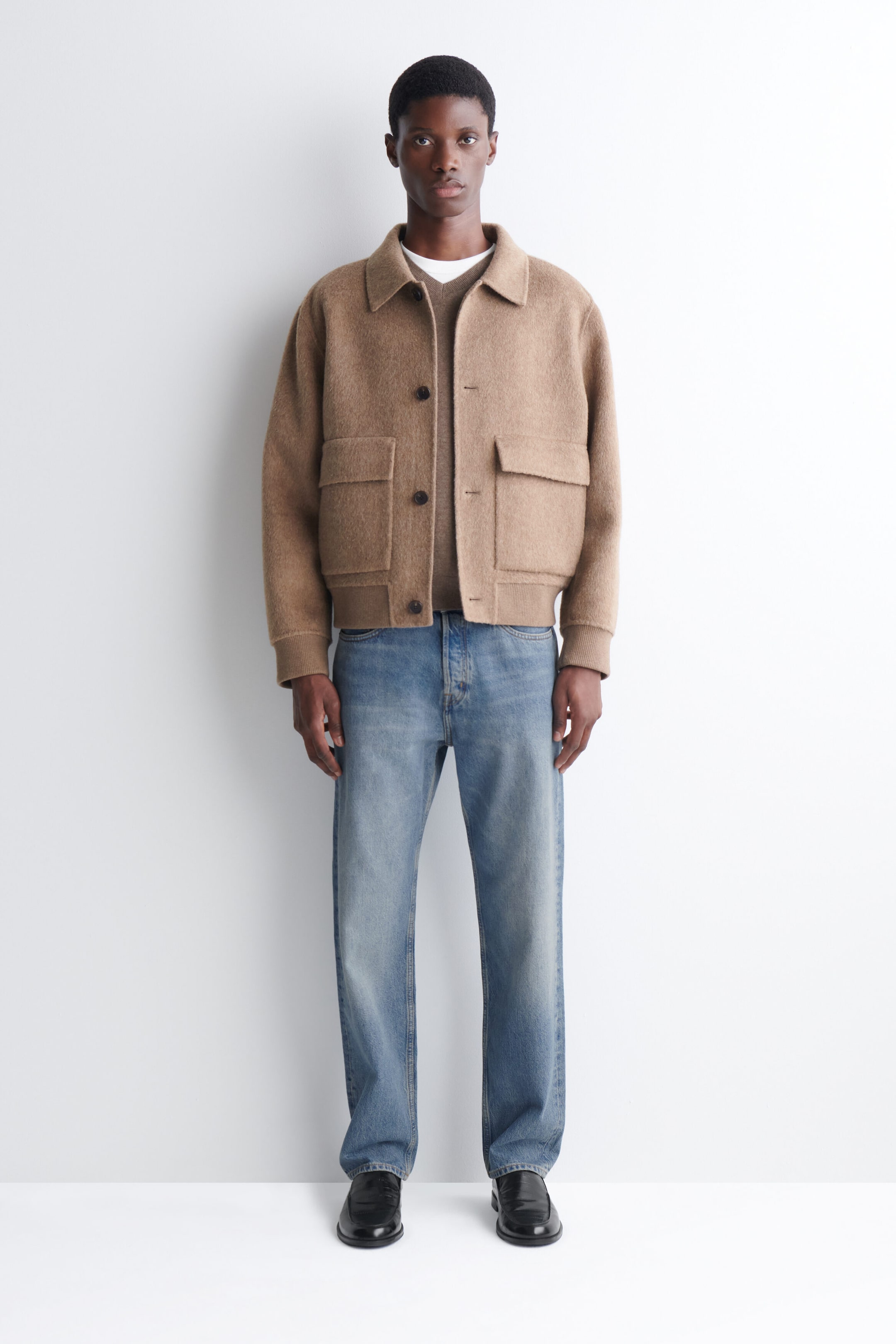 A beige double-faced blouson jacket featuring a classic collar, front button closures, and large front flap pockets is worn by the model. The jacket is layered over a matching knit sweater and styled with straight-leg blue jeans. A beige double-faced blouson jacket featuring a classic collar, front button closures, and large front flap pockets is worn by the model. The jacket is layered over a matching knit sweater and styled with straight-leg blue jeans.