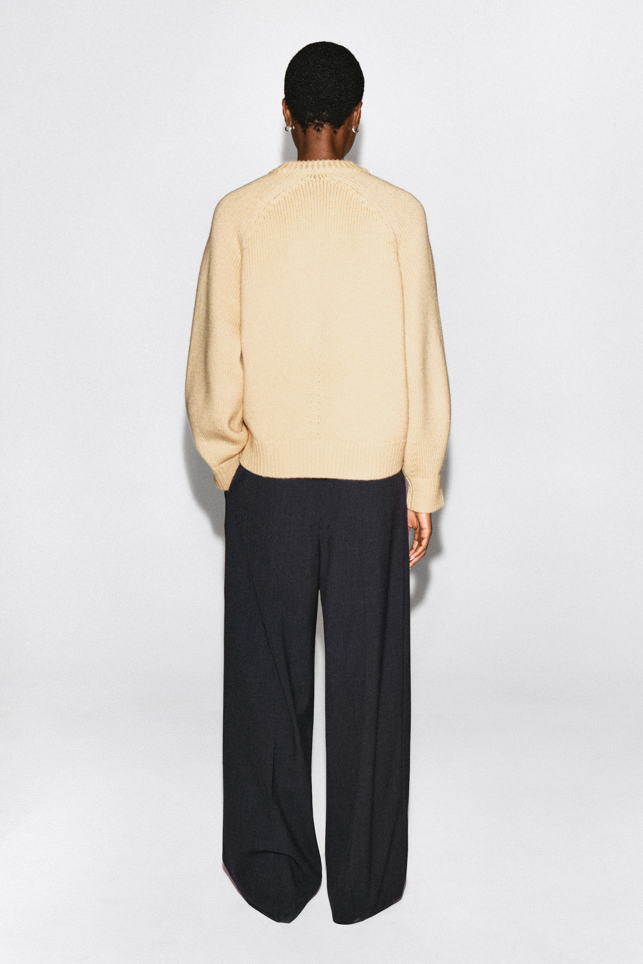 Un chandail à col rond en laine tricotée beige clair est porté par le mannequin, présenté de dos. Le chandail présente des manches raglan avec des détails côtelés au col, aux poignets et à l’ourlet, et il est porté avec un pantalon ample à jambe large foncé.