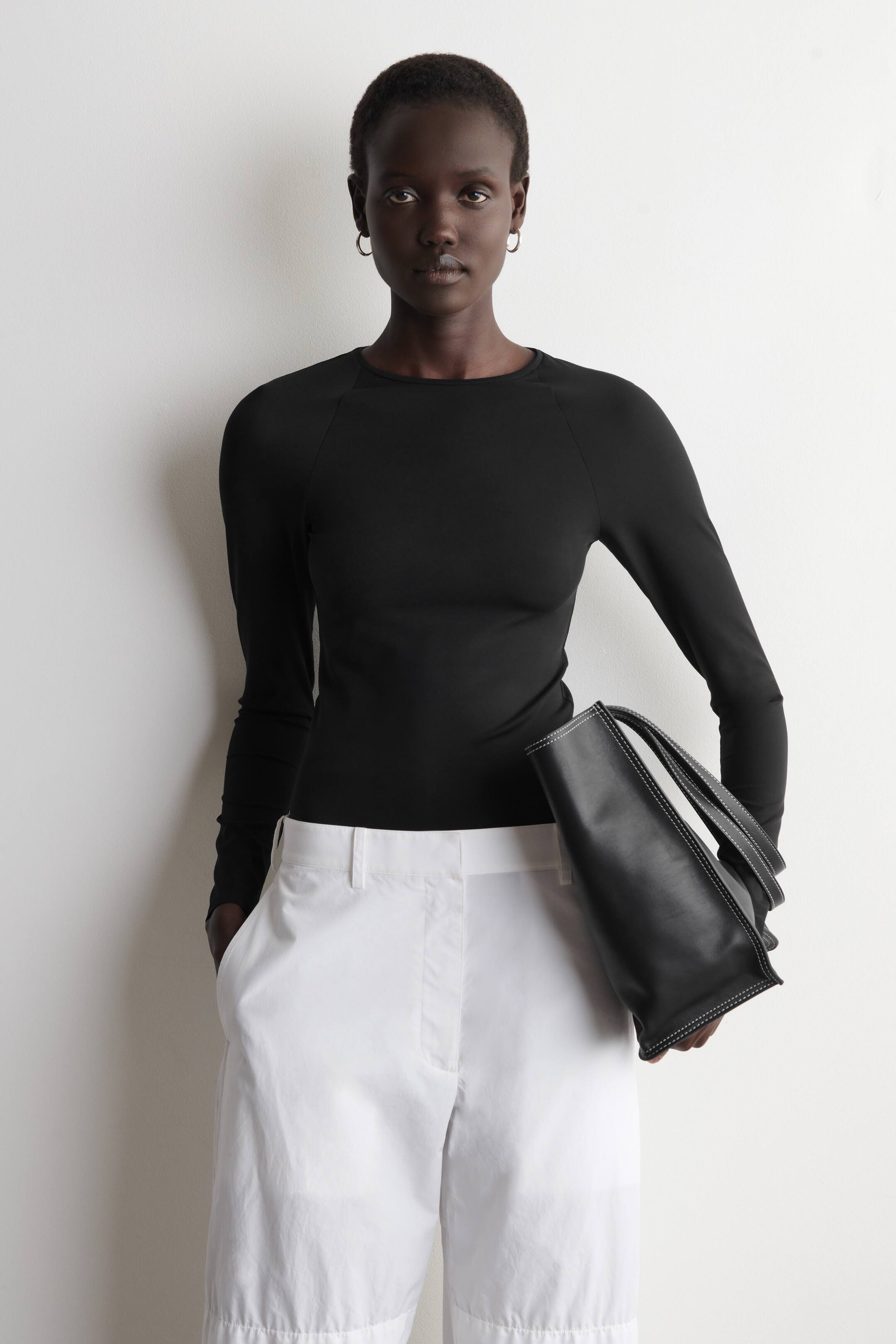 Agrandir l'image: Un haut noir à manches longues, ajusté et à col ras du cou avec des manches raglan est présenté, associé à un pantalon blanc à jambes larges et raccourcies, doté de poches latérales et de passants de ceinture. Un sac cabas noir avec des coutures contrastées blanches est tenu sous le bras.