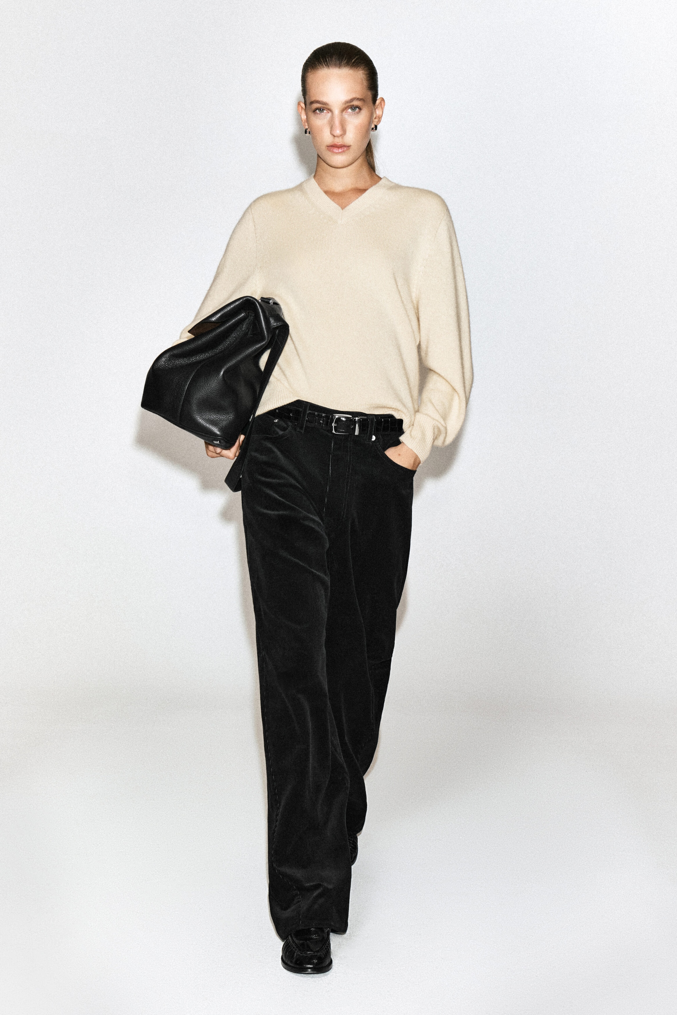 Black corduroy straight-leg jeans are worn by the model, who is walking forwards with one hand in her pocket. The jeans are styled with a black leather belt, a cream-coloured V-neck jumper, and black shoes. Black corduroy straight-leg jeans are worn by the model, who is walking forwards with one hand in her pocket. The jeans are styled with a black leather belt, a cream-coloured V-neck jumper, and black shoes.