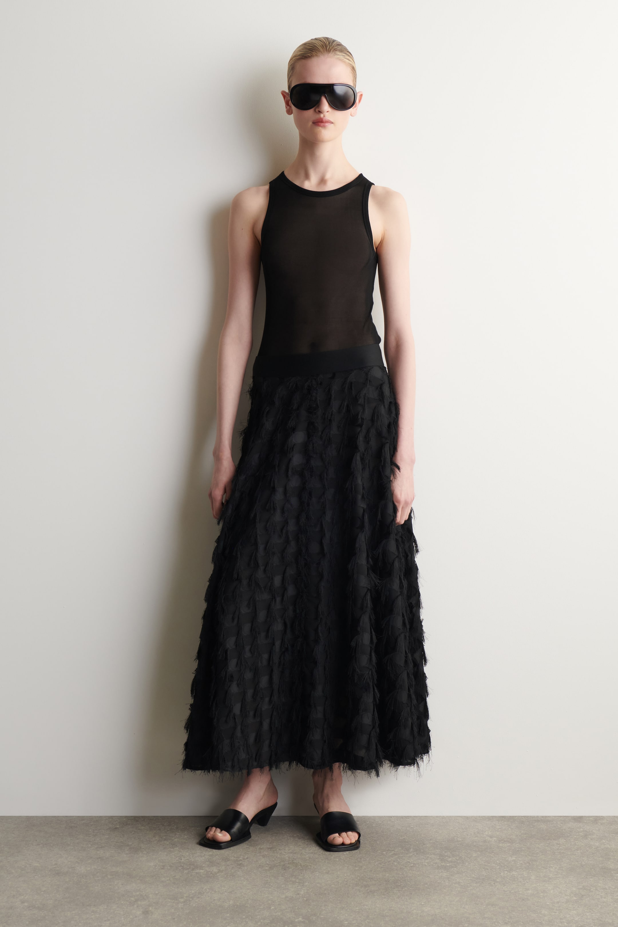 View larger image: The product is a black ensemble featuring a sheer sleeveless top with a round neckline and a midi skirt with an all-over textured, feathery design. It is styled with oversized black shield sunglasses and black open-toed mules with block heels.