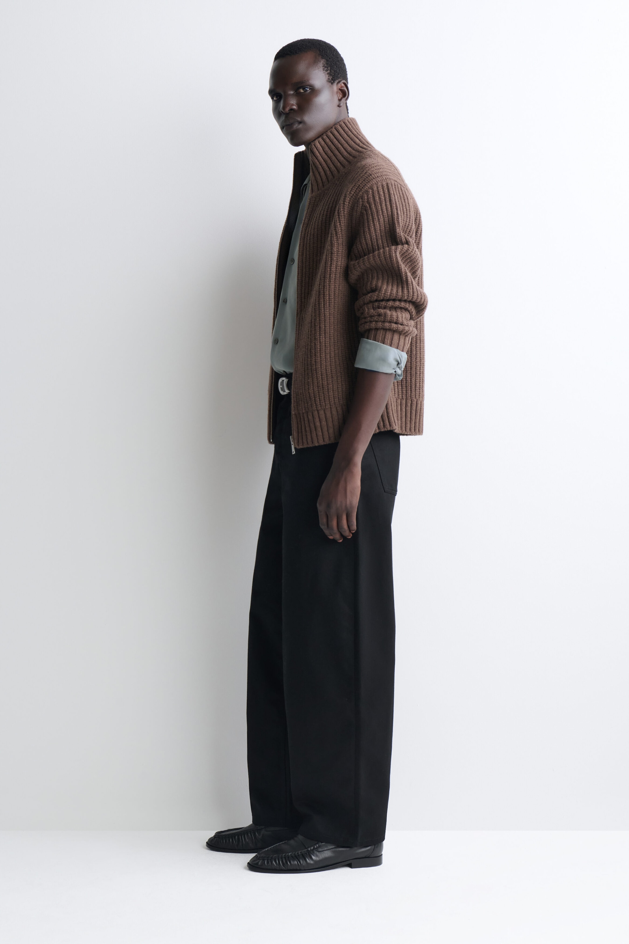 A brown, heavyweight ribbed-knit zip-up cardigan with a high funnel neck is facing left, styled over a light gray button-down shirt and wide-leg black trousers. The cardigan features a thick vertical ribbed texture, with its sleeves pushed up to reveal the folded cuffs of the shirt underneath. A brown, heavyweight ribbed-knit zip-up cardigan with a high funnel neck is facing left, styled over a light gray button-down shirt and wide-leg black trousers. The cardigan features a thick vertical ribbed texture, with its sleeves pushed up to reveal the folded cuffs of the shirt underneath.